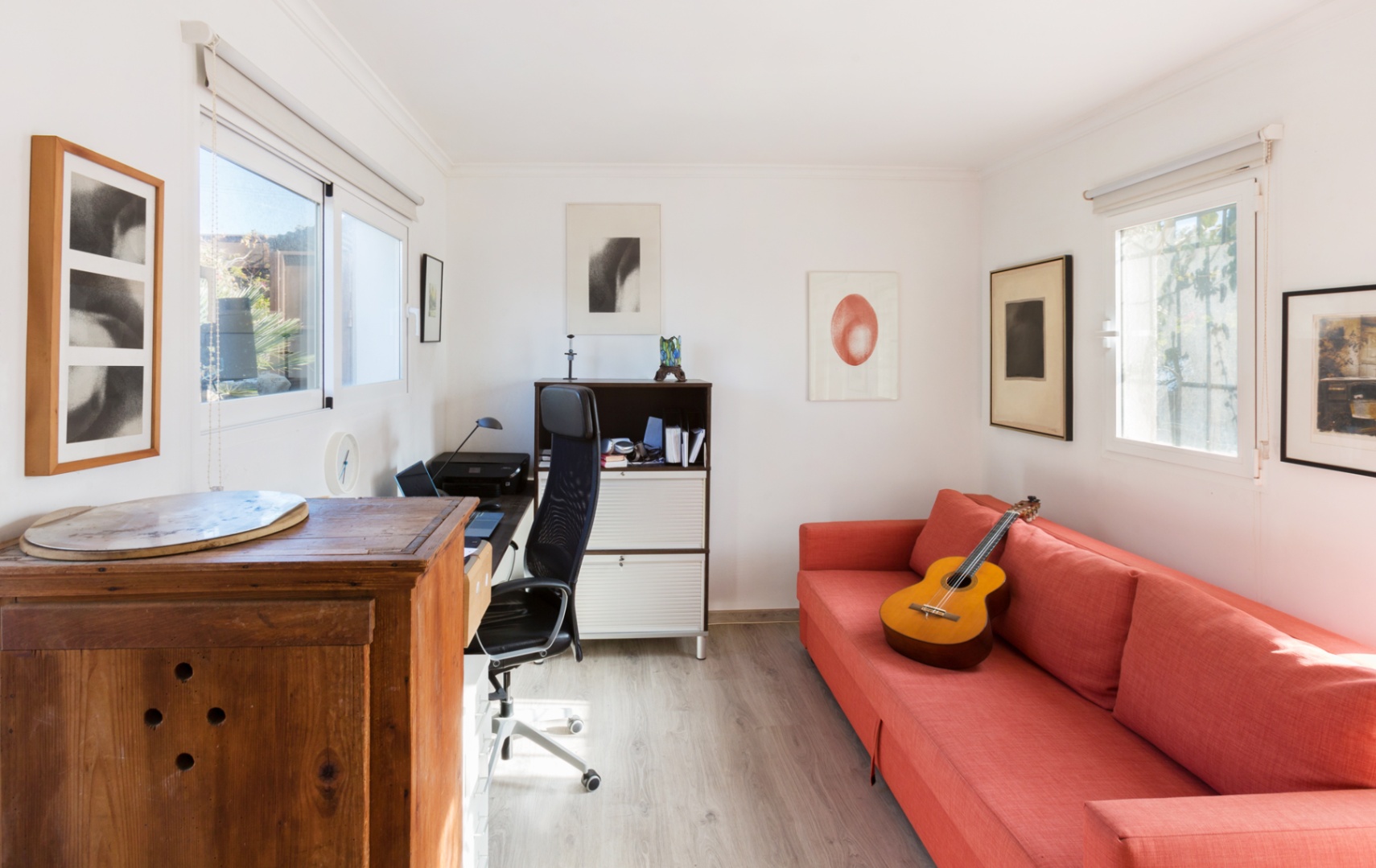 Helles Arbeitszimmer mit hellem Holzboden, rotem Sofa, ergonomischem Arbeitsplatz und natürlichem Lichteinfall durch zwei Fenster mit Rollos.