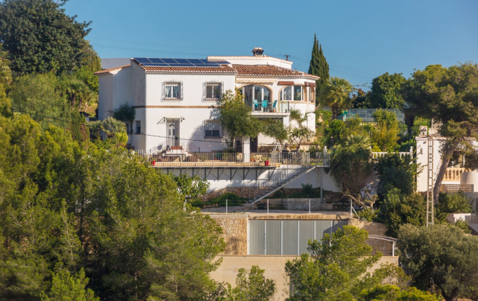 Mehrstöckige mediterrane Villa mit weißer Fassade, Solaranlage auf dem Dach und Terrassen, eingebettet in eine grüne Hügellandschaft mit Natursteinmauern.