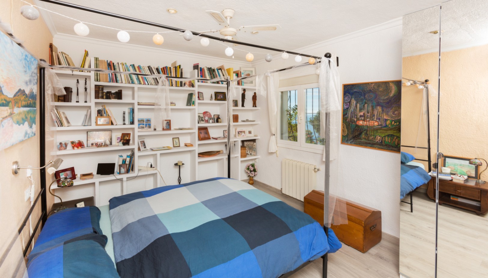Schlafzimmer mit schwarzem Himmelbett, eingebauten weißen Regalen und großem Spiegelschrank. Inklusive Deckenventilator und Fenster mit Blick auf die Küstenlandschaft.
