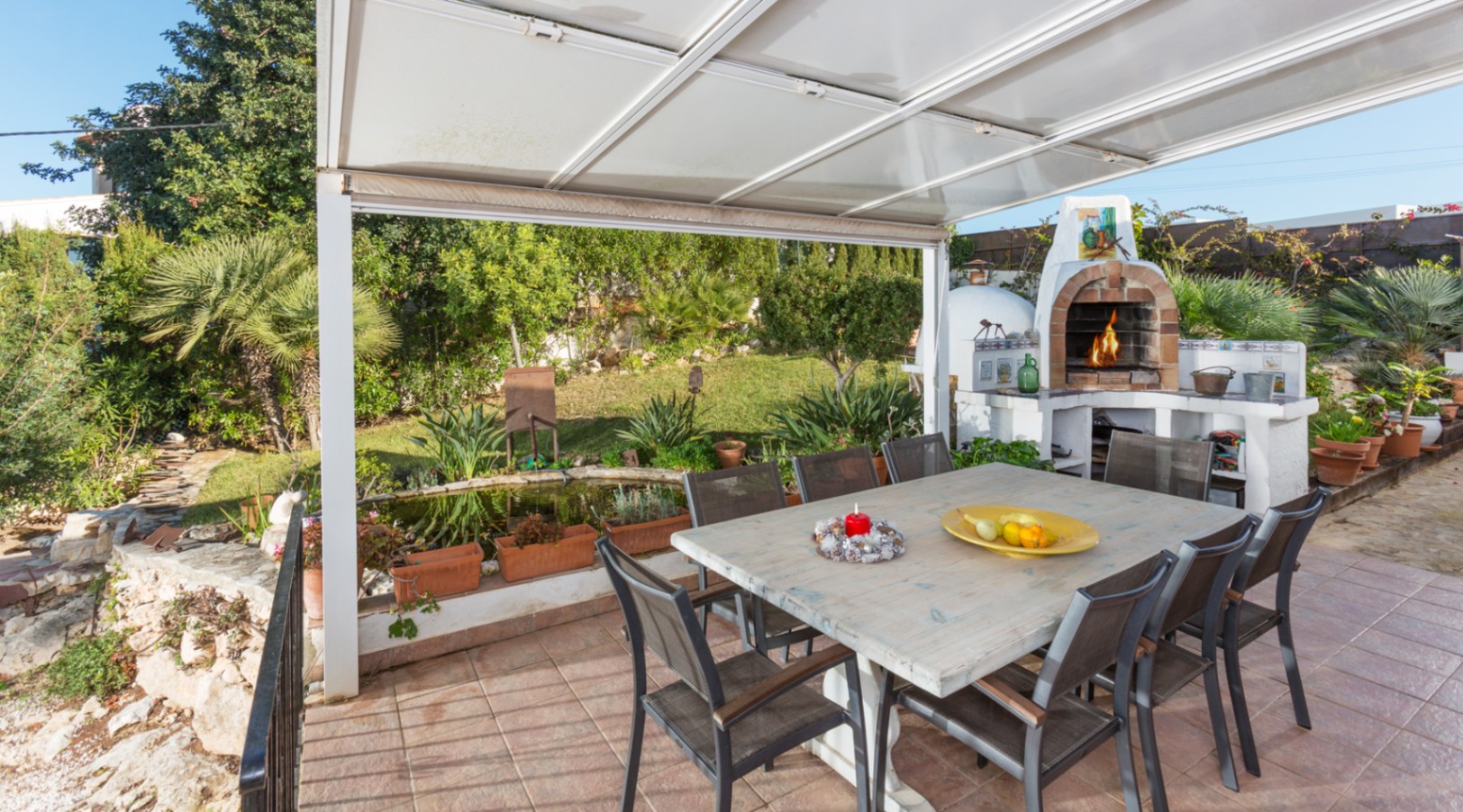 Überdachte Terrasse mit großem Holztisch, traditionellem gemauertem Holzofen und Blick auf den mediterranen Garten mit Teich.