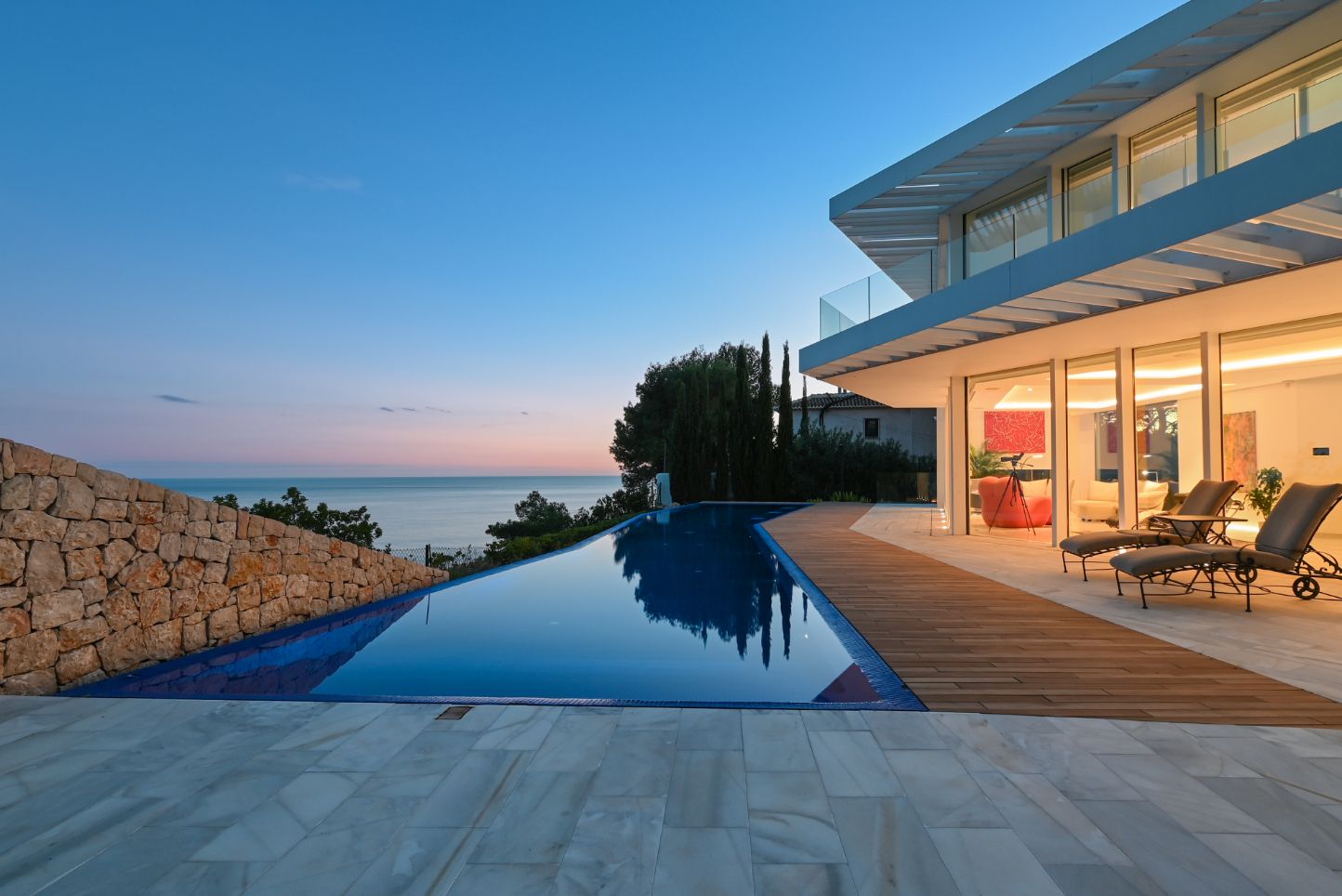 Infinity-Pool mit Holzdeck und Steinpflasterung mit Blick auf das Meer. Moderne Villa mit großen Glasfronten und Natursteinmauer bei Dämmerung.
