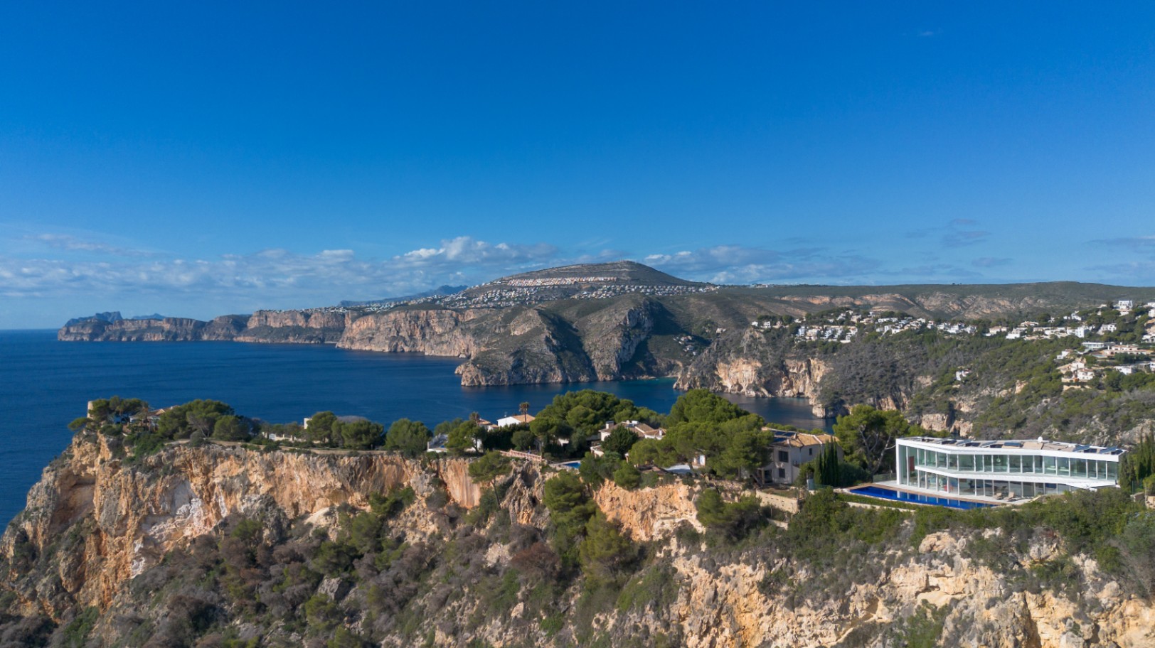 Luftaufnahme einer modernen Villa mit Glasfront auf einer Klippe, mit linearem Pool und Panoramablick auf die mediterrane Küstenlandschaft.