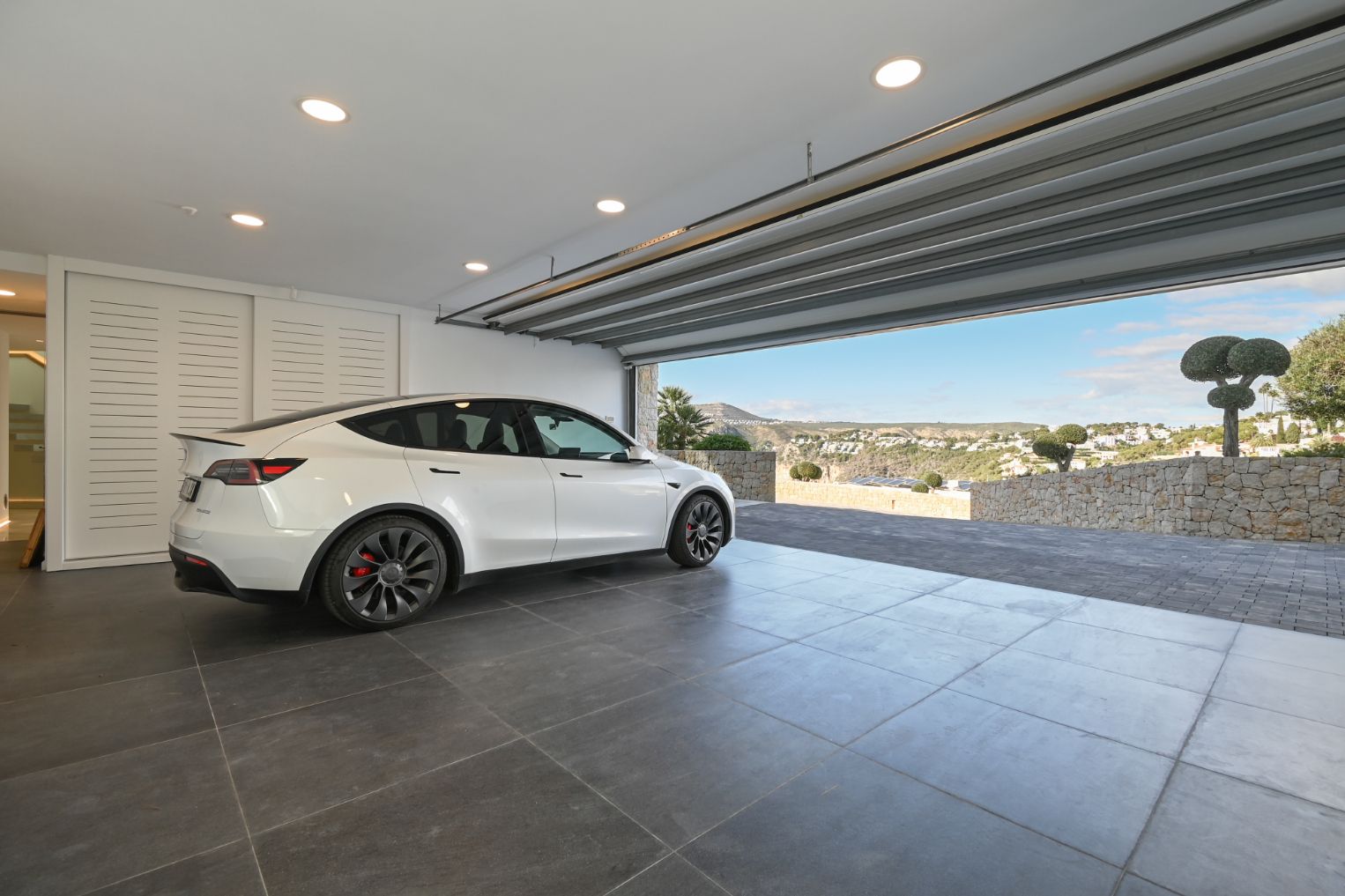 Moderne Garage mit großformatigen grauen Bodenfliesen, Deckeneinbaustrahlern und freiem Blick auf das Tal über die gepflasterte Steineinfahrt.