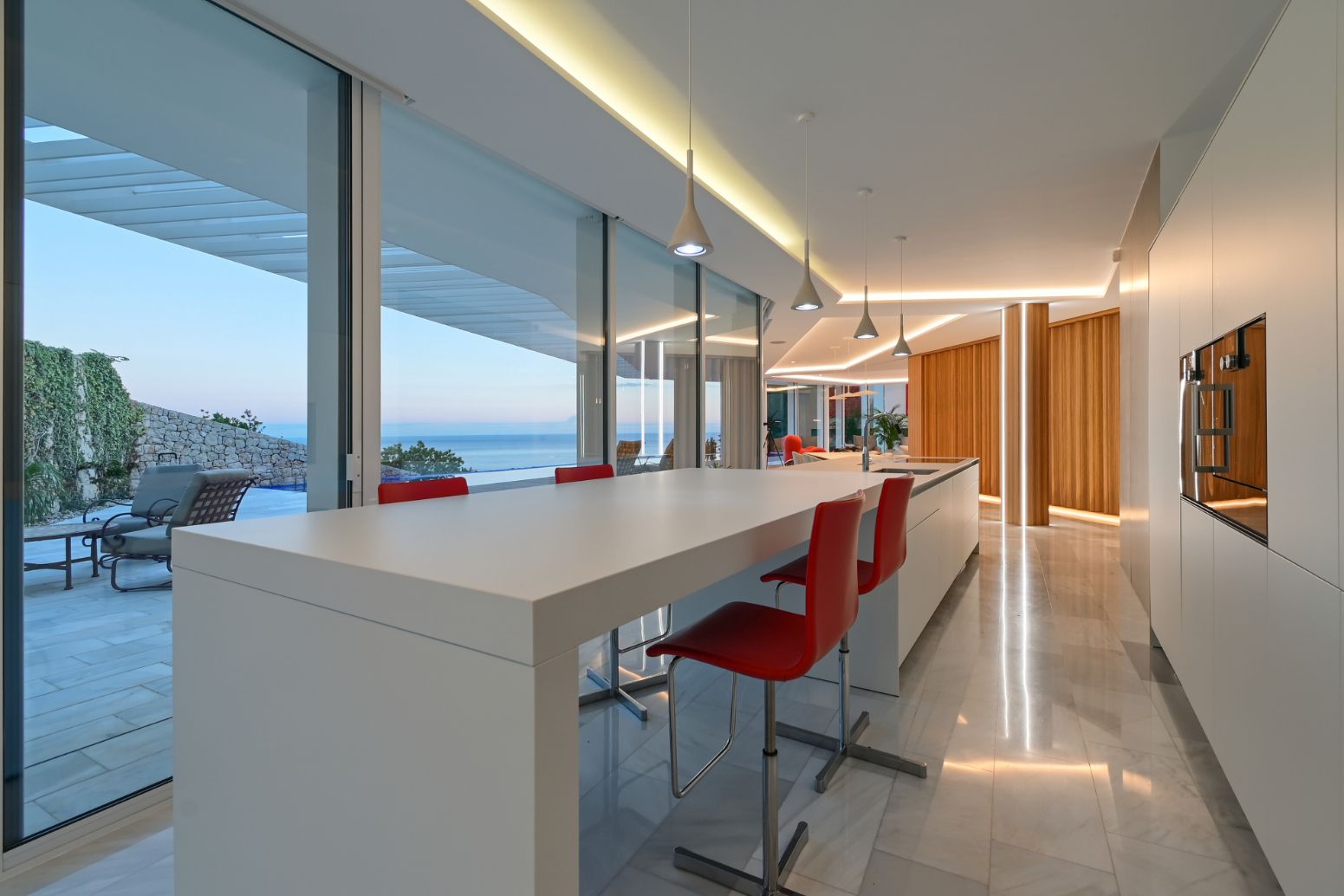 Moderne Küche mit weißer minimalistischer Insel, roten Barhockern, bodentiefen Fenstern mit Meerblick und integrierter LED-Beleuchtung.