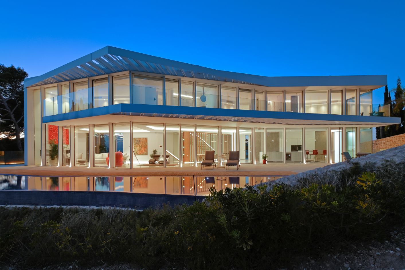 Moderne zweistöckige Villa mit bodentiefen Glasfassaden, weitläufiger Terrasse mit Pool und integrierter Innenbeleuchtung in der Abenddämmerung.