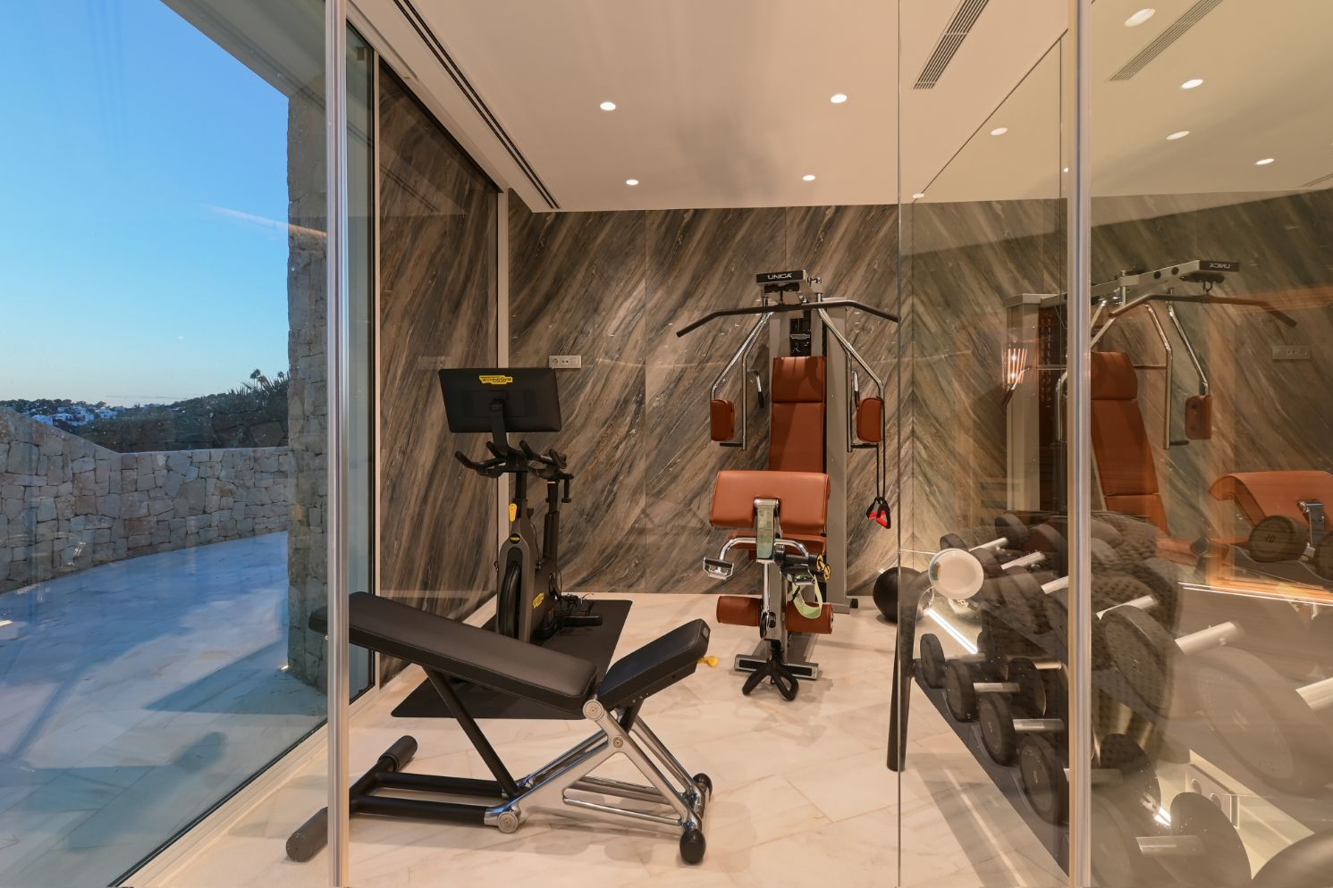 Moderner Fitnessraum mit Technogym-Geräten, marmorierten Wandpaneelen und bodentiefen Glasfronten mit Blick auf eine Außenterrasse aus Naturstein.
