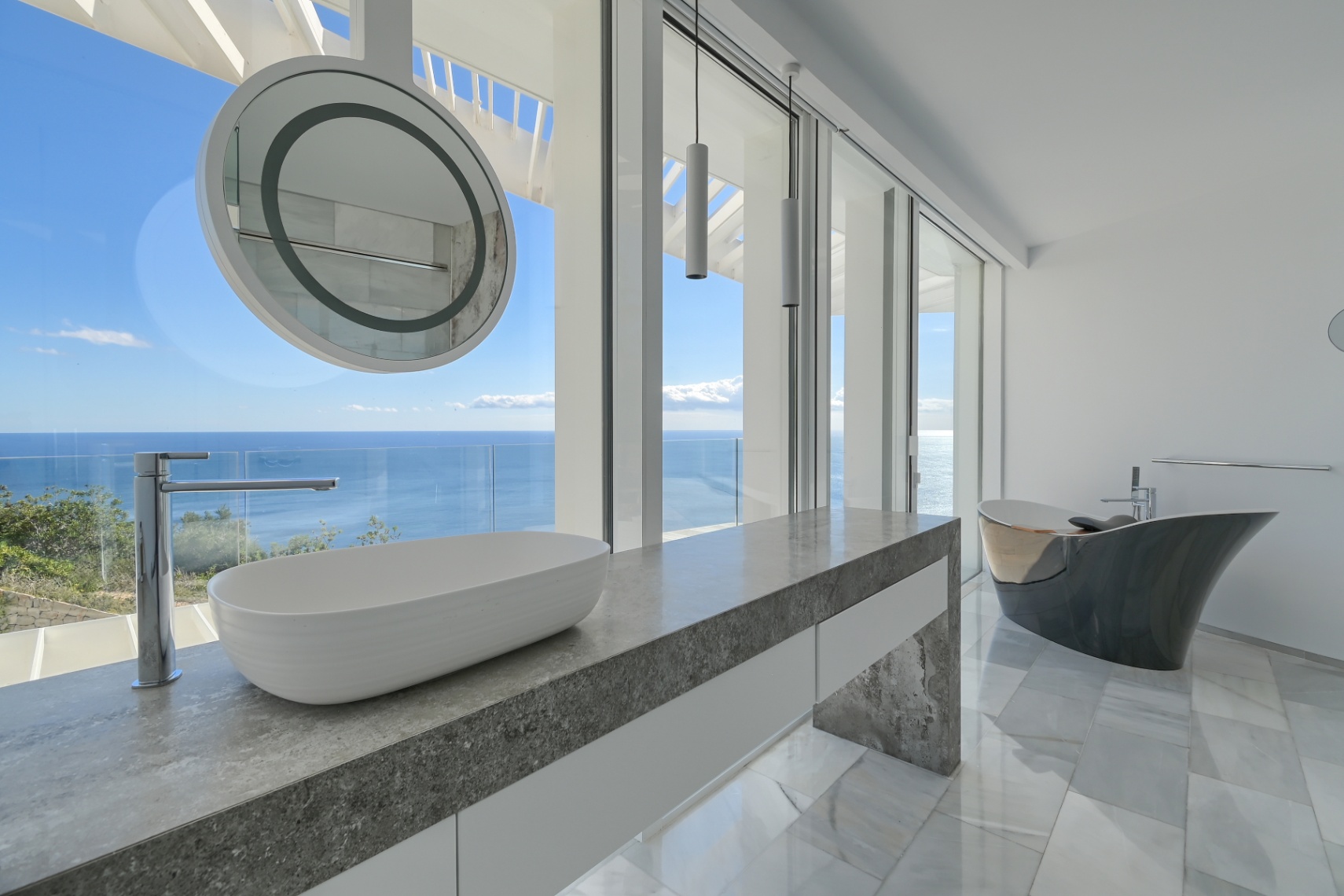 Modernes Badezimmer mit Waschtisch aus grauem Stein, Aufsatzwaschbecken und freistehender Design-Badewanne sowie bodentiefen Fenstern mit Meerblick.