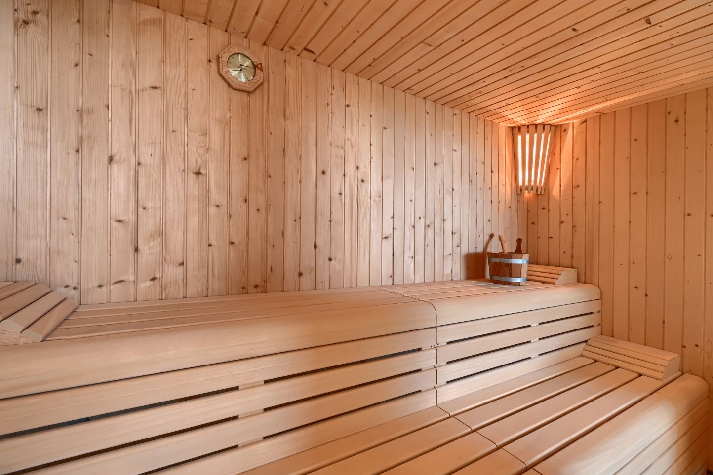 Sauna-Innenraum mit heller Holzvertäfelung, mehrstufigen Sitzbänken, einer Eckleuchte und traditionellem Zubehör wie Holzeimer und Kelle.