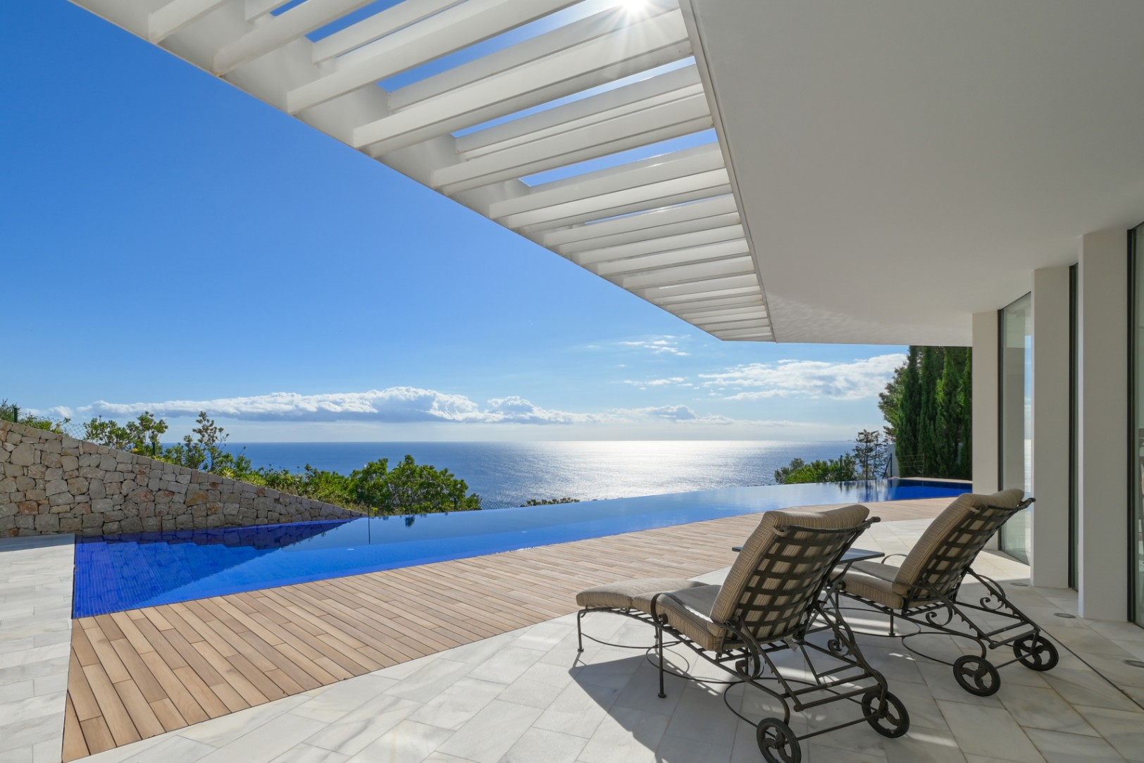 Terrasse mit Infinity-Pool und Panoramablick auf das Meer, Holzdeck und Marmorboden unter einer weißen Pergola mit eleganten Sonnenliegen.
