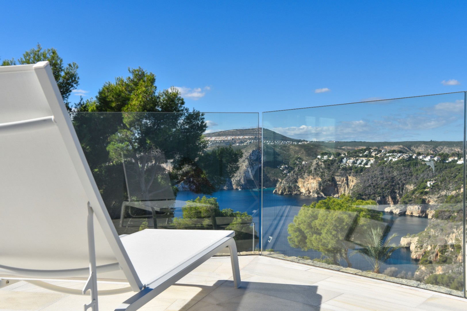 Terrasse mit weißer Sonnenliege und Glasgeländer, mit Panoramablick auf eine Mittelmeerbucht und bewaldete Küstenfelsen bei Tageslicht.