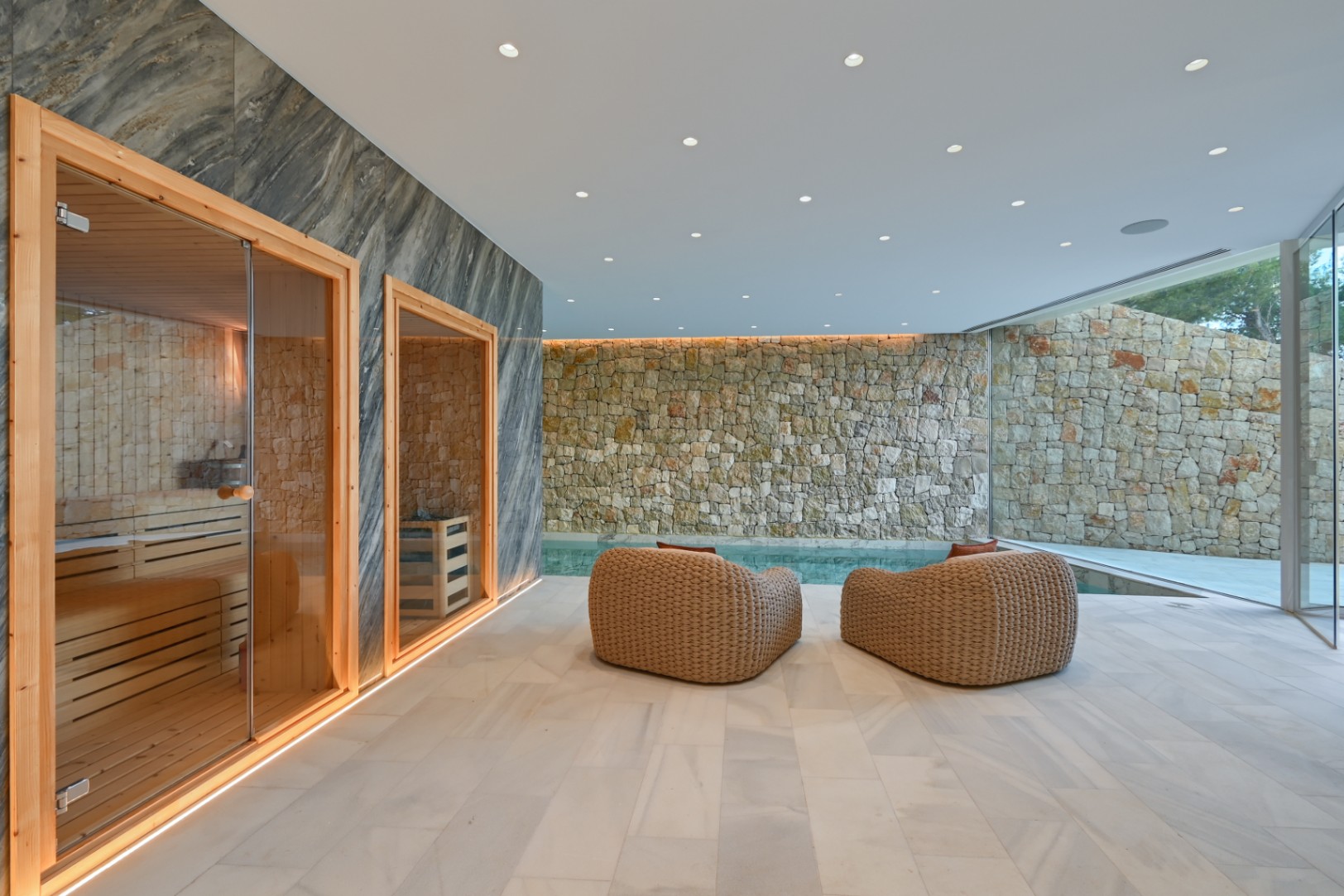 Wellnessbereich im Innenbereich mit Holzsauna, geflochtenen Loungesesseln und einer rustikalen Steinwand neben dem privaten Pool.