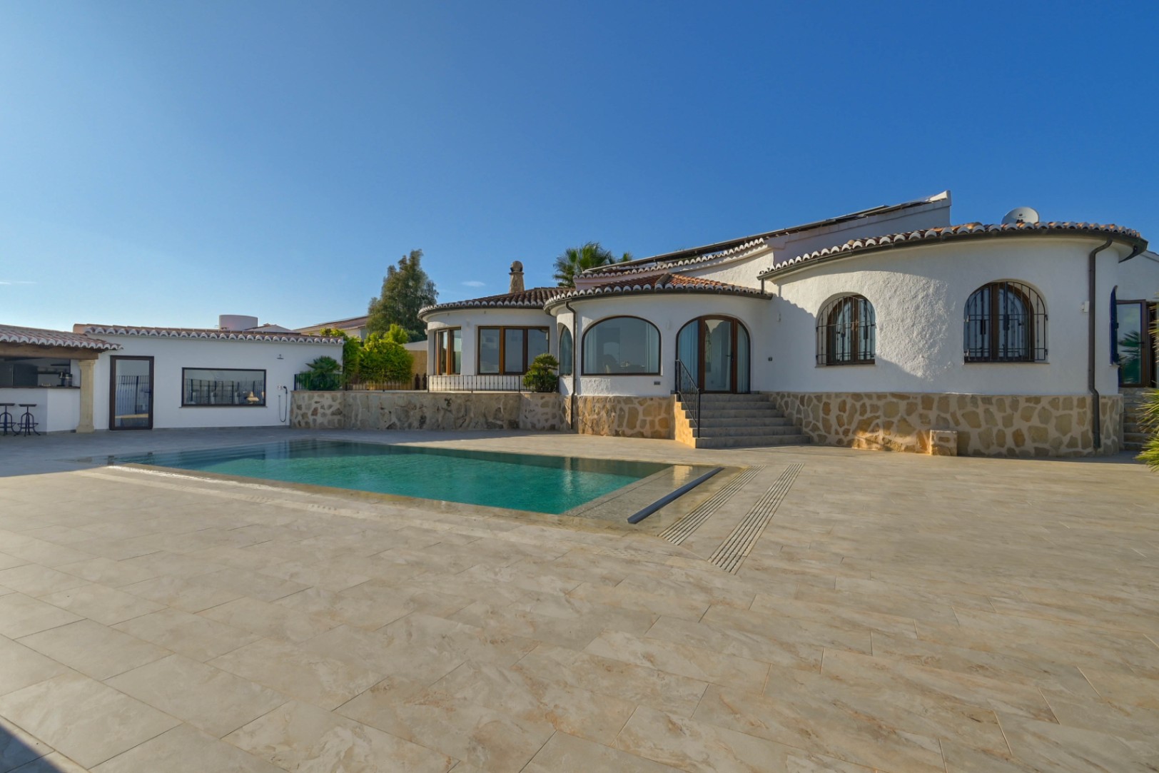 Außenansicht einer mediterranen Villa mit rechteckigem Pool, Natursteinsockel und großzügiger Fliesenterrasse bei klarem Himmel.