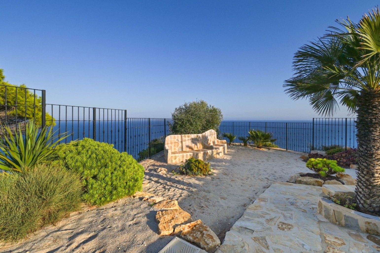 Aussichtsterrasse mit gemauerter Steinbank, schwarzem Metallzaun und Panoramablick auf das Meer, umgeben von mediterranen Pflanzen und Palmen.