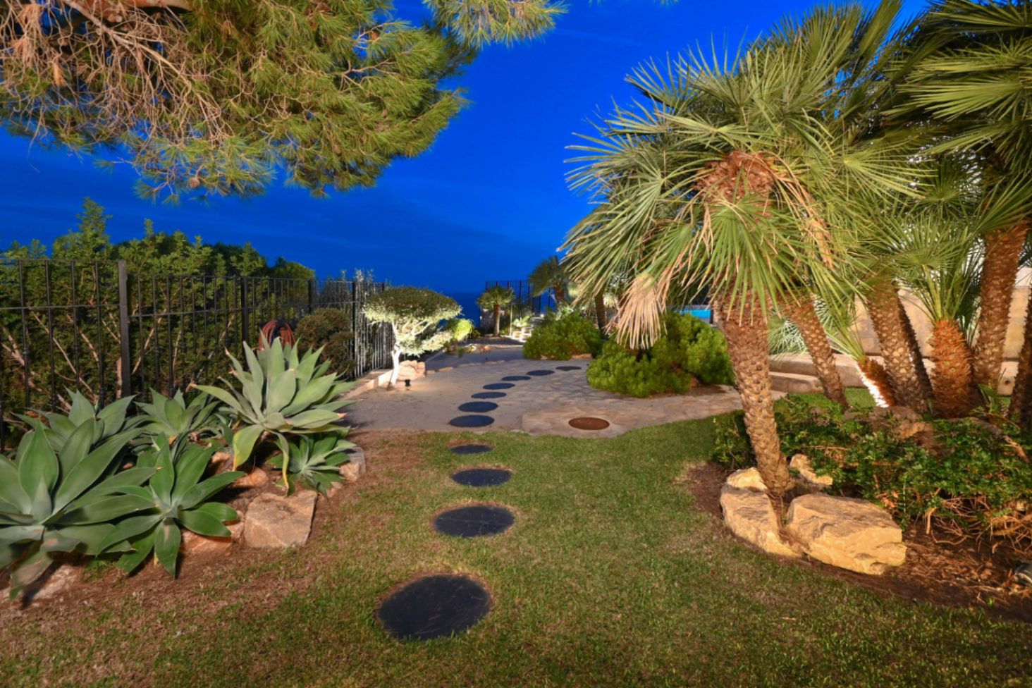 Gartenpfad mit kreisförmigen Trittsteinen, Palmen, Agaven und integrierter Außenbeleuchtung in einem mediterranen Garten bei Dämmerung.