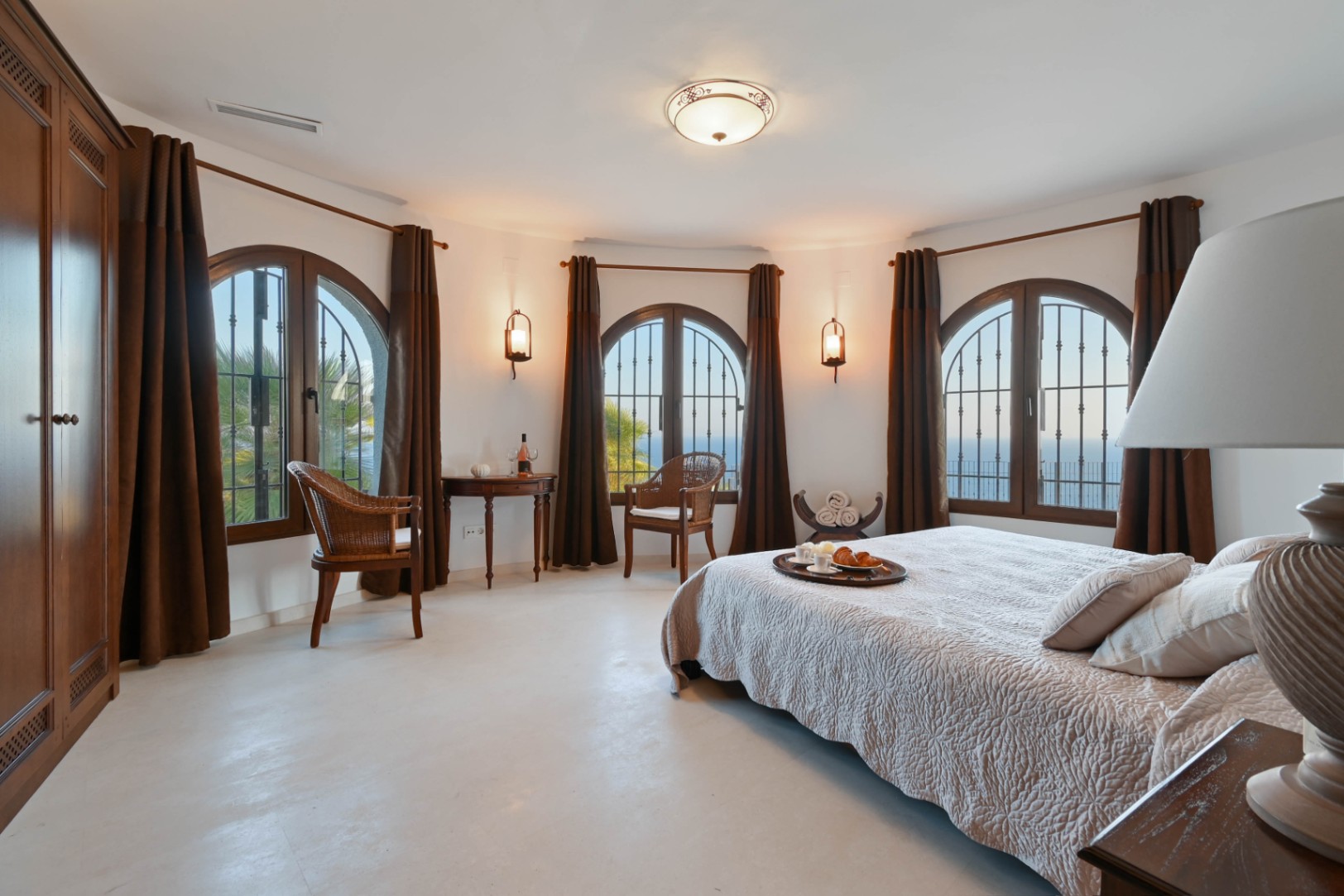 Großzügiges Schlafzimmer mit geschwungener Wand, drei Rundbogenfenstern aus dunklem Holz, Korbsesseln und weitem Meerblick.