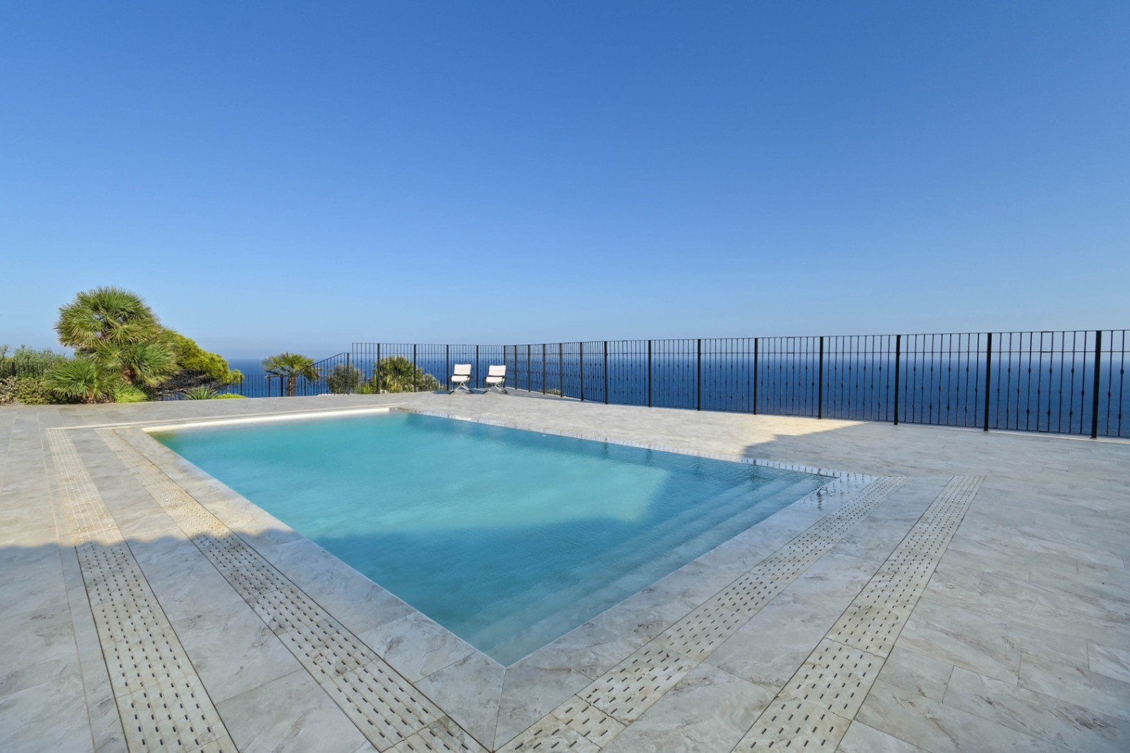 Rechteckiger Swimmingpool mit integrierter Treppe auf einer hellen Steinterrasse, eingefasst von einem schwarzen Metallgeländer mit weitem Meerblick.