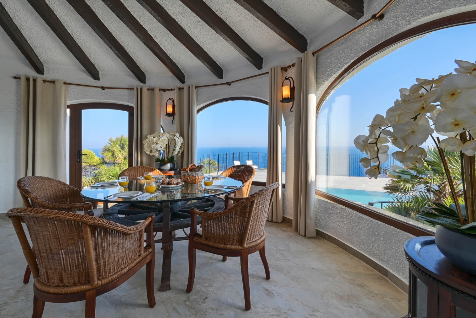 Rundes Esszimmer mit Glastisch auf einem Wagenrad und Korbstühlen unter einer Holzbalkendecke, mit weitem Blick auf das Meer durch bogenförmige Fensterfronten.
