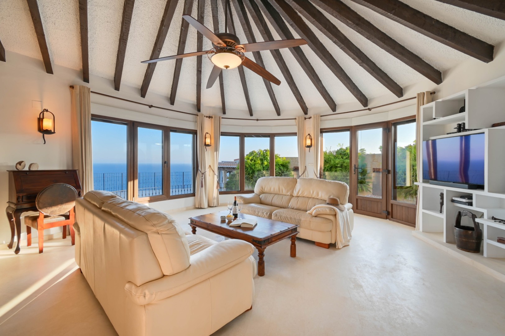 Rundes Wohnzimmer mit gewölbter Balkendecke, Ledersofas und großen Panoramafenstern, die einen weiten Blick auf das Meer bieten.