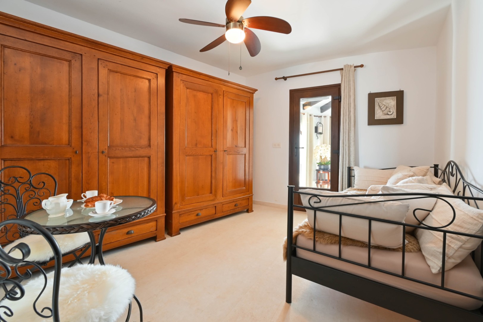 Schlafzimmer mit zwei großen rustikalen Holzschränken, einem schwarzen Schmiedeeisen-Tagesbett und Bistrotisch. Inklusive Deckenventilator und Glastür nach draußen.