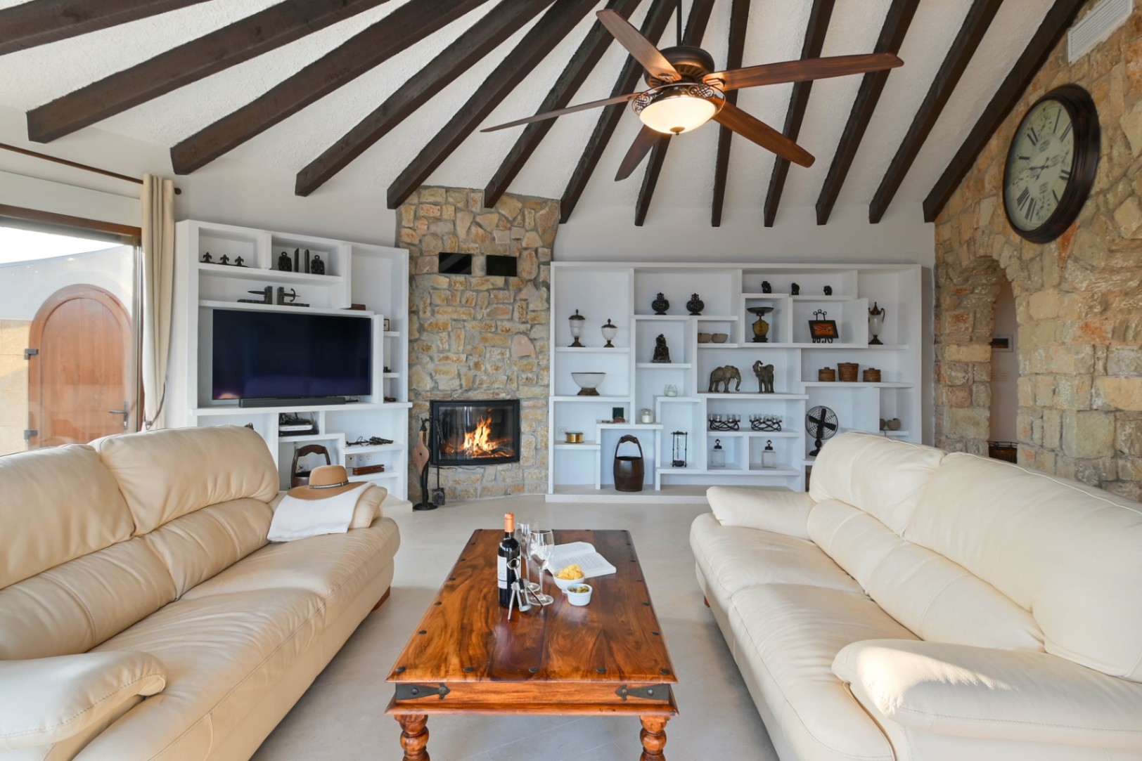 Wohnzimmer mit Holzbalkendecke, Natursteinkamin und integrierten weißen Regalen. Ausgestattet mit cremefarbenen Ledersofas und einem rustikalen Couchtisch aus Holz.