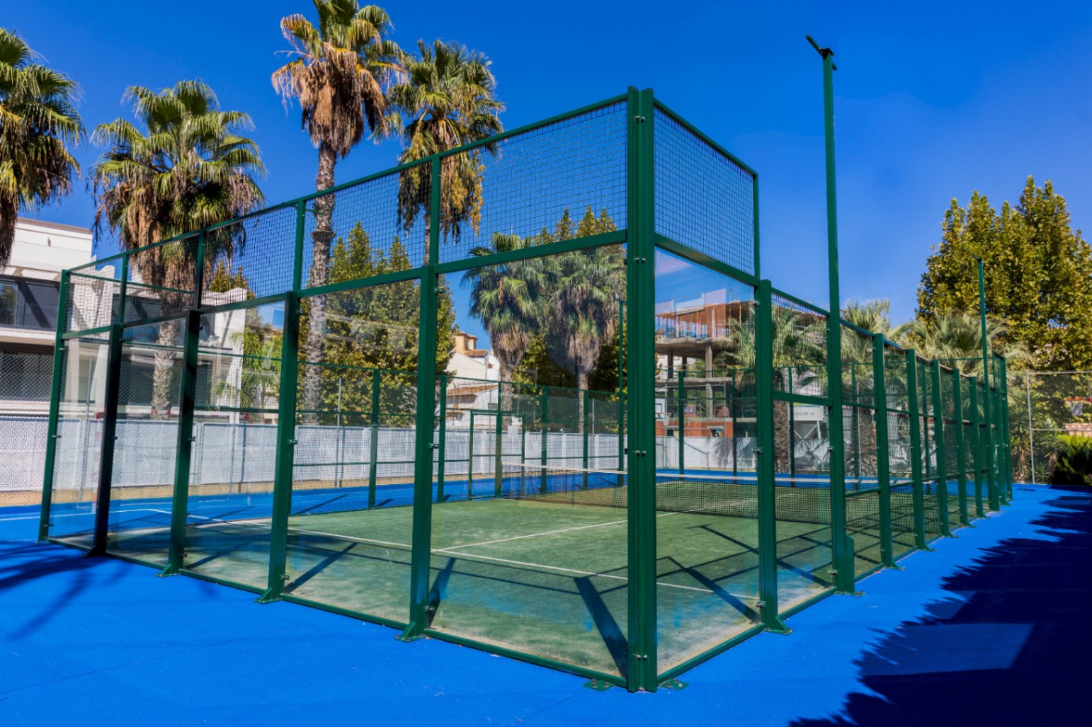 Außen-Paddle-Tennisplatz mit grünem Kunstrasen, Glaswänden und grünem Metallrahmen, umgeben von Palmen und einem blauen Bodenbelag.