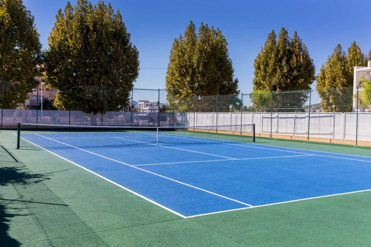 Außen-Tennis-Hartplatz mit blauem Spielfeld und grünem Rand, umgeben von einem hohen Schutzzaun und altem Baumbestand unter klarem Himmel.