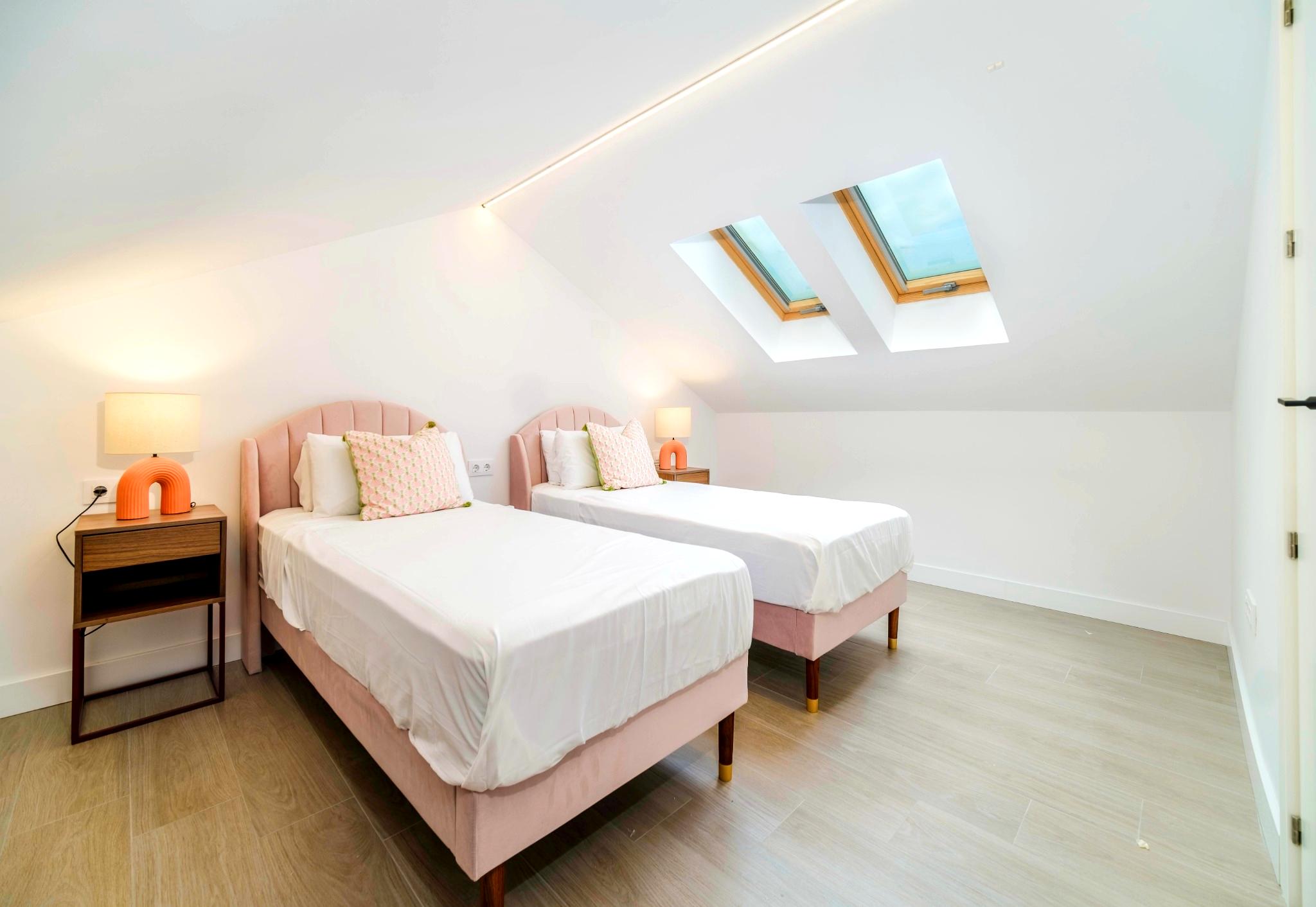 Dachgeschoss-Schlafzimmer mit zwei Einzelbetten aus rosa Samt, doppelten Dachfenstern und integrierter LED-Lichtleiste an der schrägen Decke.