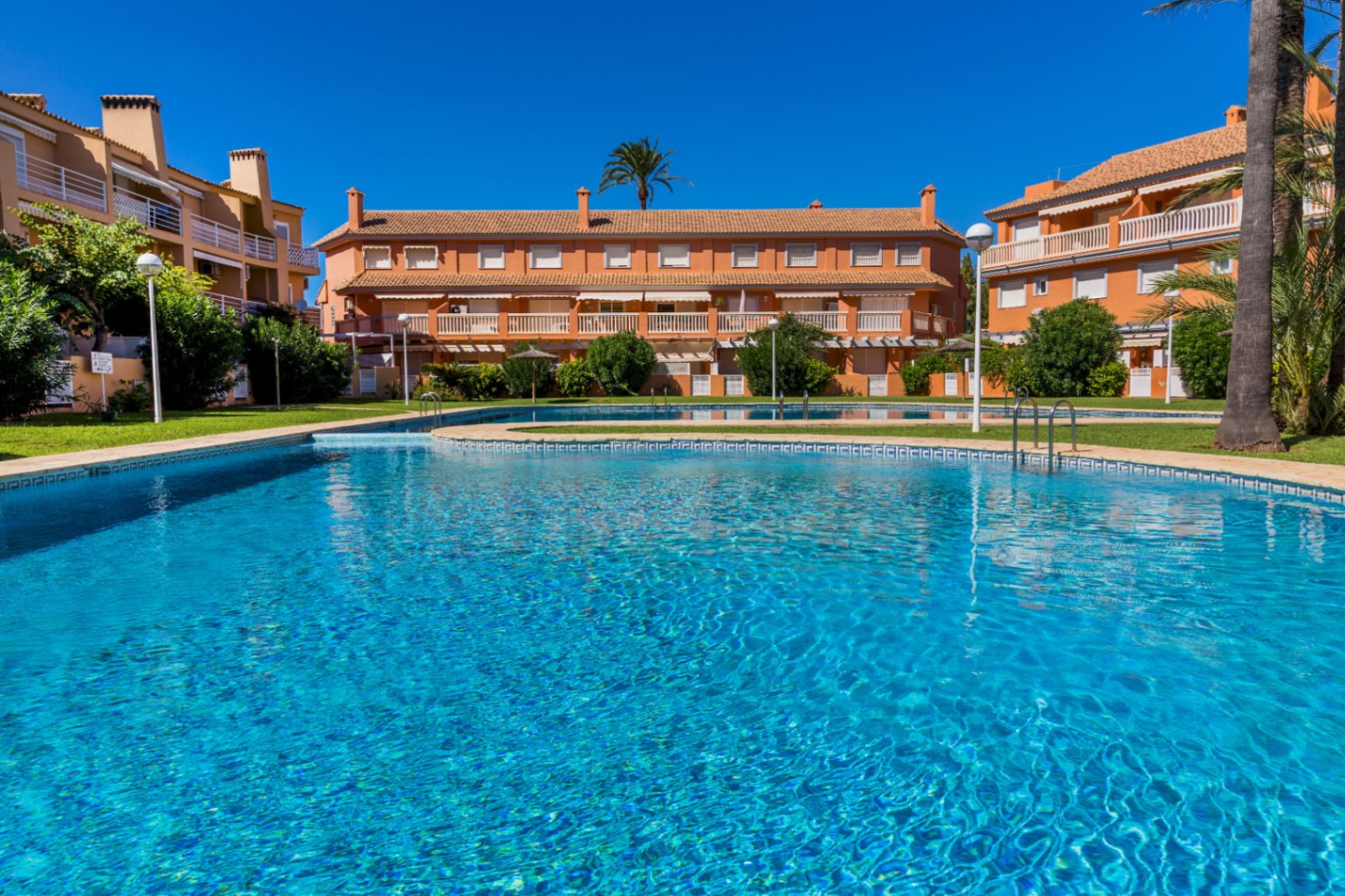 Großer Gemeinschaftspool mit blauen Mosaikfliesen, umgeben von gepflegten Rasenflächen und terrakottafarbenen Wohngebäuden mit Balkonen.