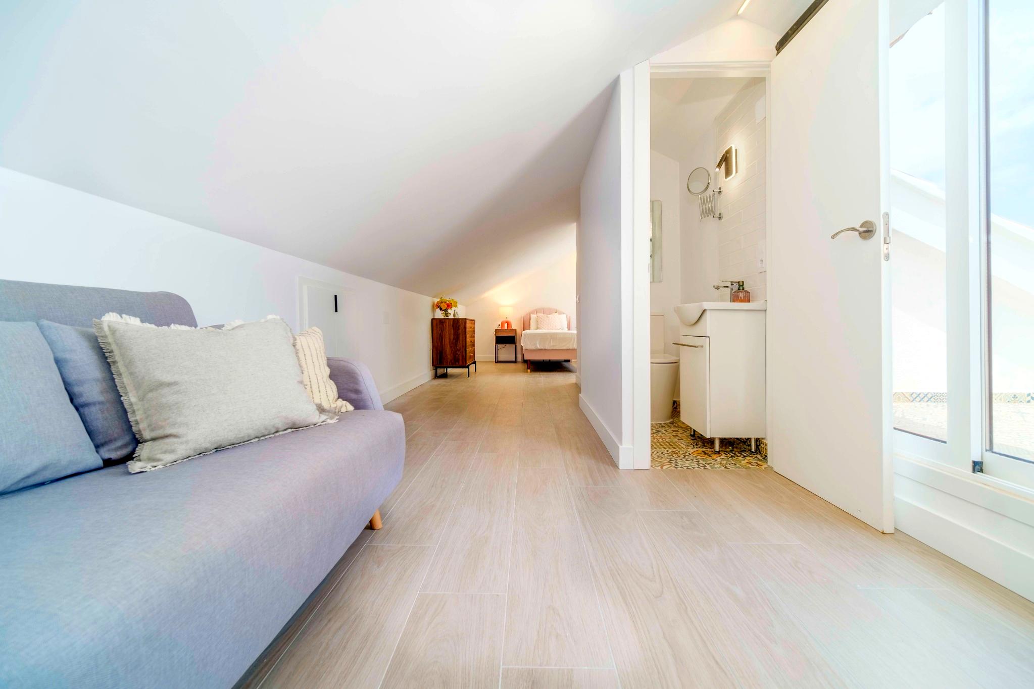 Helle Dachgeschoss-Suite mit weißen Dachschrägen, Bodenfliesen in Holzoptik, Wohnbereich und angrenzendem Badezimmer mit gemusterten Fliesen.