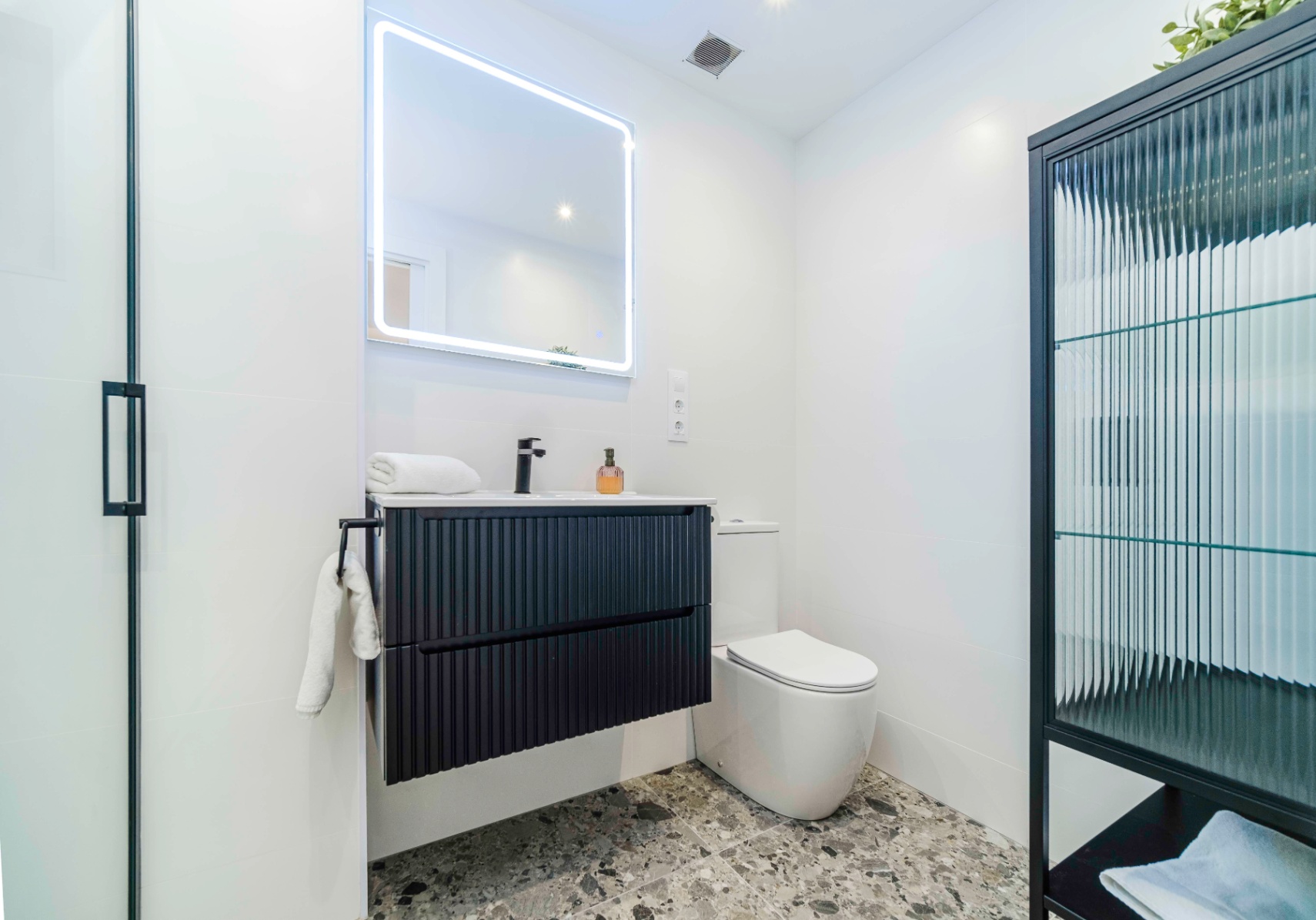 Modernes Badezimmer mit schwarzem, geriffeltem Waschtisch, hintergrundbeleuchtetem LED-Spiegel und grauem Terrazzoboden.