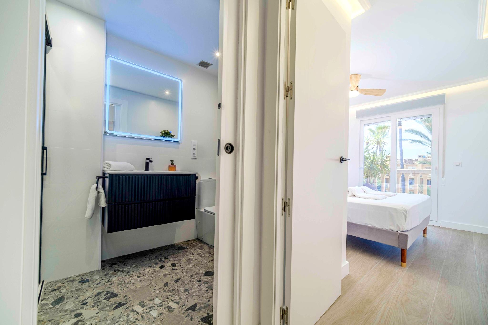 Modernes Badezimmer mit Terrazzoboden und schwarzem Waschtisch, angrenzend an ein helles Schlafzimmer mit Balkon und Blick auf Palmen.