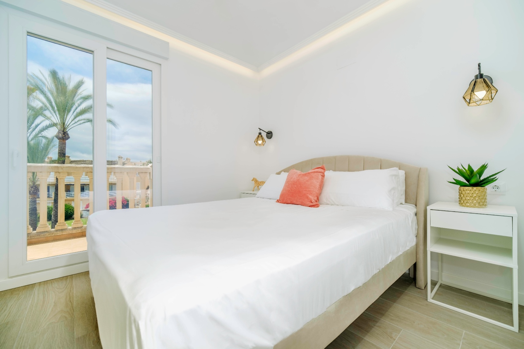 Modernes Schlafzimmer mit gepolstertem Kopfteil, geometrischen Wandleuchten und Glasschiebetür zur Terrasse mit Blick auf Palmen.