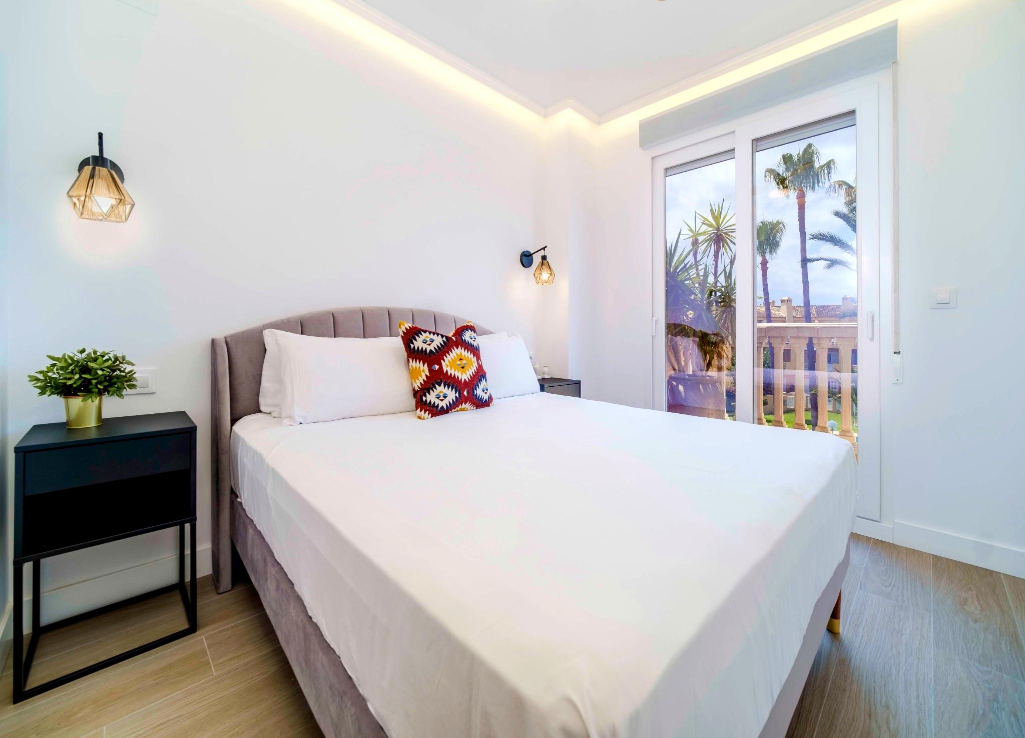 Modernes Schlafzimmer mit Samtkopfteil, geometrischen Wandleuchten und indirekter Deckenbeleuchtung. Große Glasschiebetüren führen zum Balkon mit Blick auf Palmen.