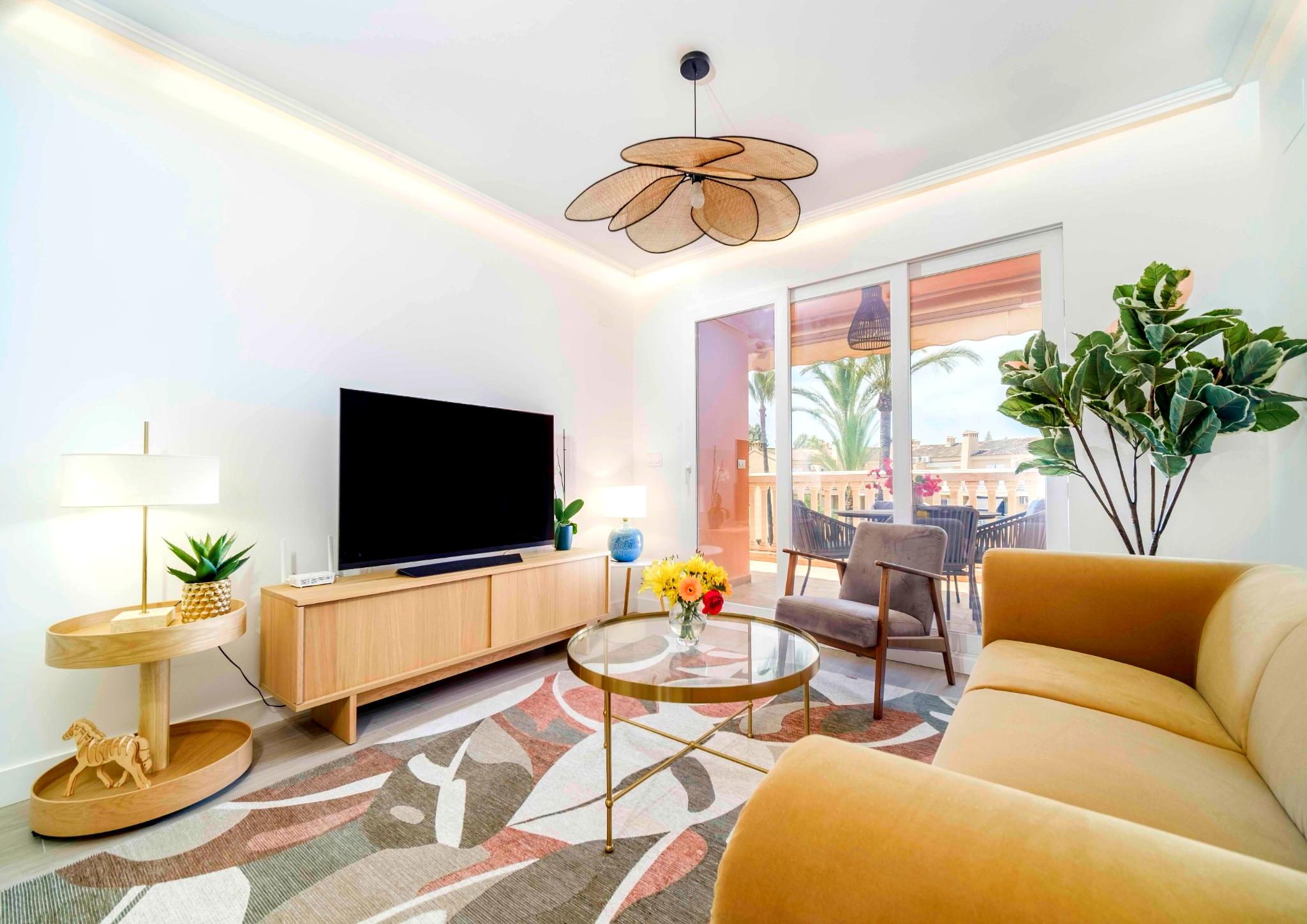 Modernes Wohnzimmer mit gelbem Sofa, einer Deckenleuchte in Blütenform und Glasschiebetüren, die auf eine Terrasse mit Palmenblick führen.