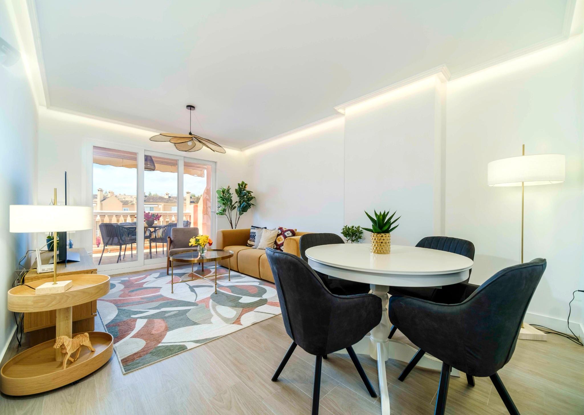 Offener Wohn- und Essbereich mit senfgelbem Sofa, rundem Esstisch und indirekter LED-Deckenbeleuchtung. Schiebetüren führen zur möblierten Terrasse.