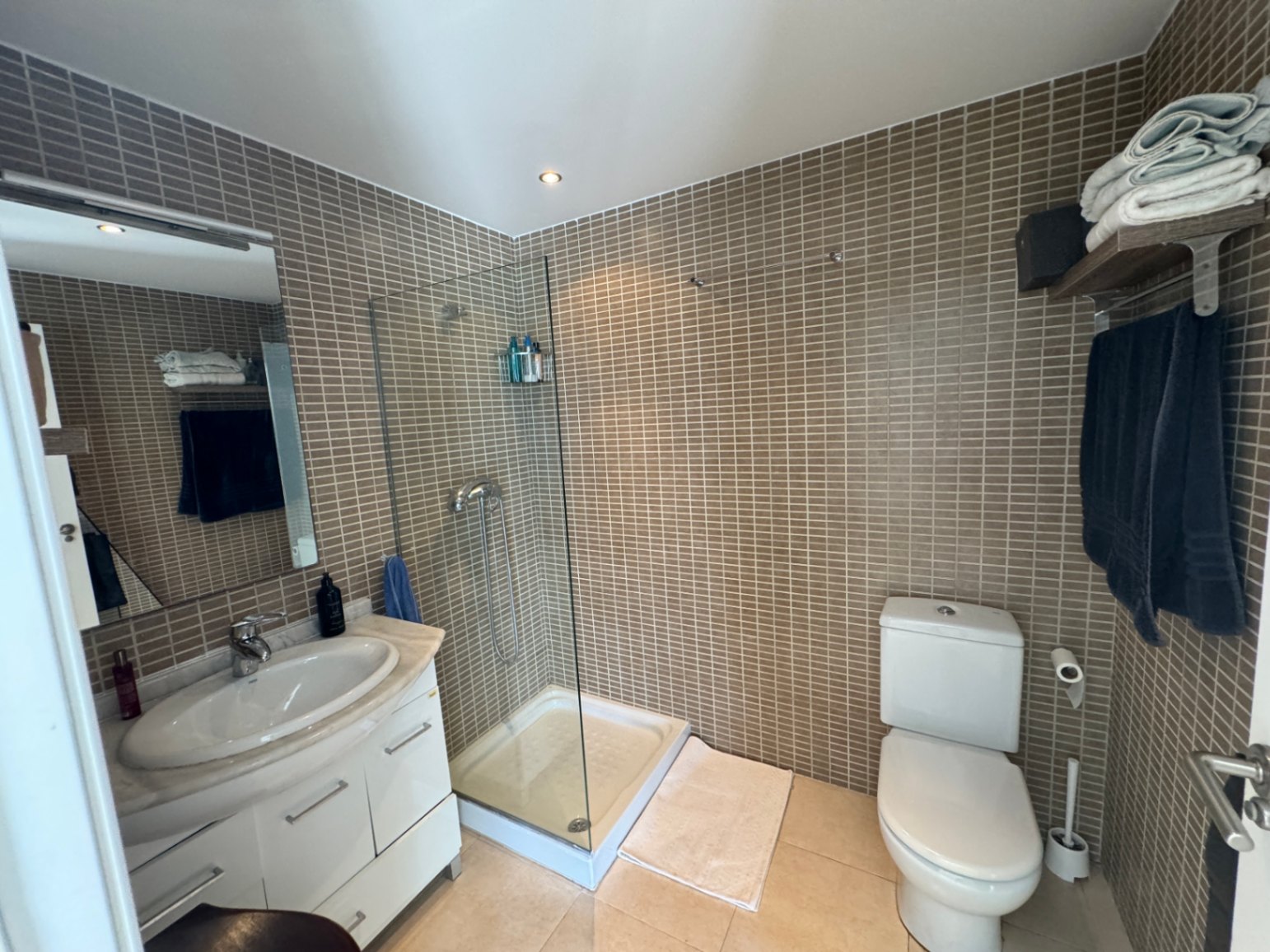 Badezimmer mit Glasduschkabine, braunen Mosaikwänden und weißem Waschtisch mit Marmorplatte sowie Deckeneinbaustrahlern.