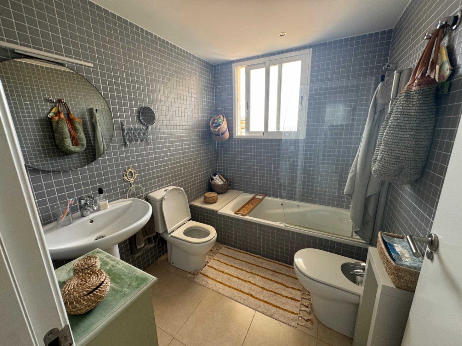 Badezimmer mit grauen Mosaikfliesen, weißem Säulenwaschbecken, Rundspiegel und einer Badewanne mit Glastrennwand sowie natürlichem Lichteinfall durch das Fenster.