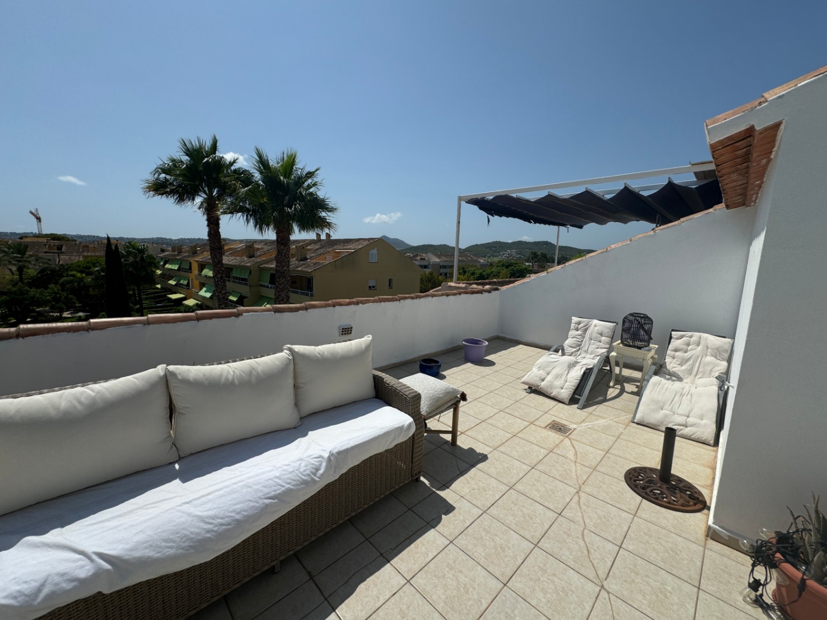 Dachterrasse mit Fliesenboden, Korbsofa, Sonnenliegen und ausziehbarer Markise. Blick auf Palmen und die umliegende Wohngegend.