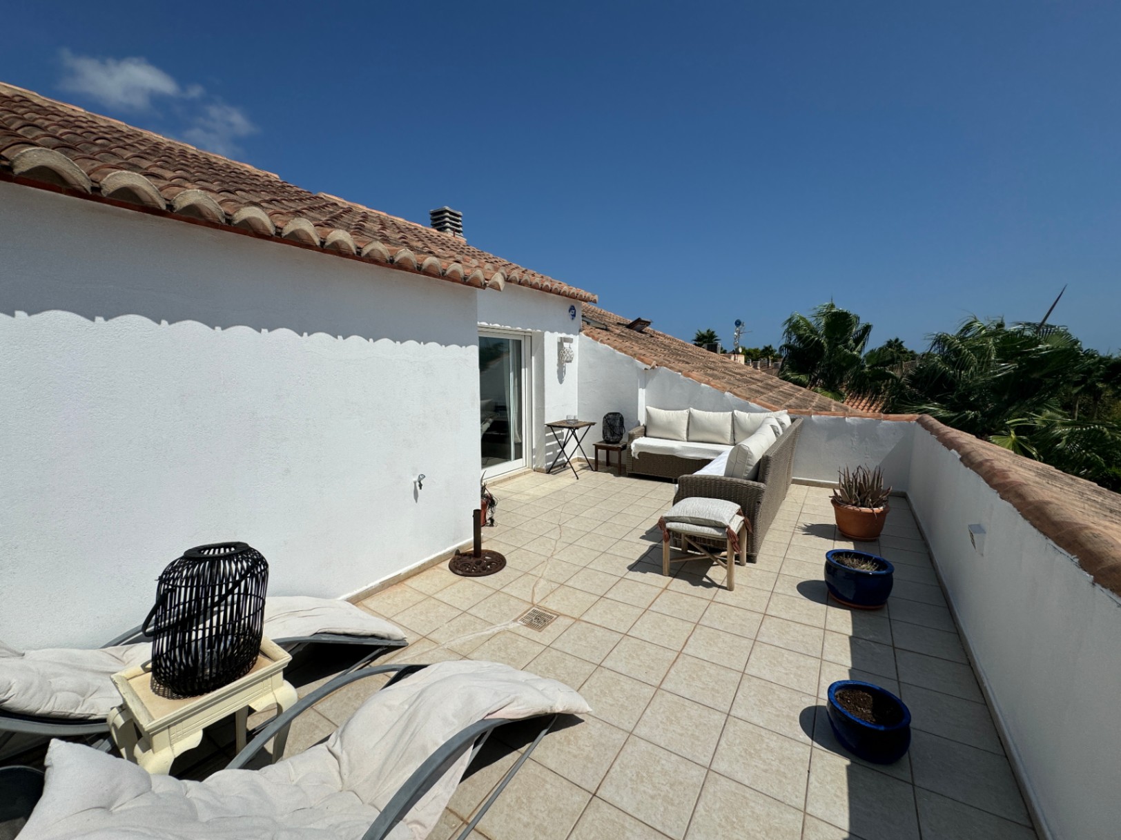 Dachterrasse mit weißen Putzwänden, Terrakotta-Ziegeldach, Rattan-Sitzgruppe und Fliesenboden mit Blick auf Palmen.