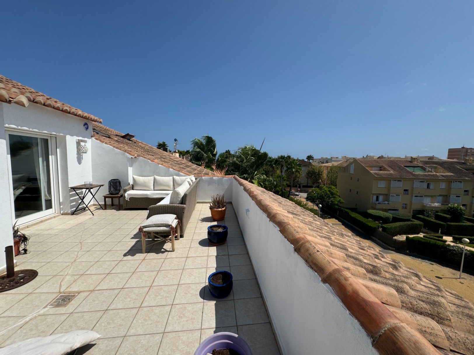Großzügige Dachterrasse mit hellen Bodenfliesen, einer L-förmigen Rattan-Sitzgruppe und Blick auf die umliegende Wohngegend mit Palmen und Ziegeldächern.