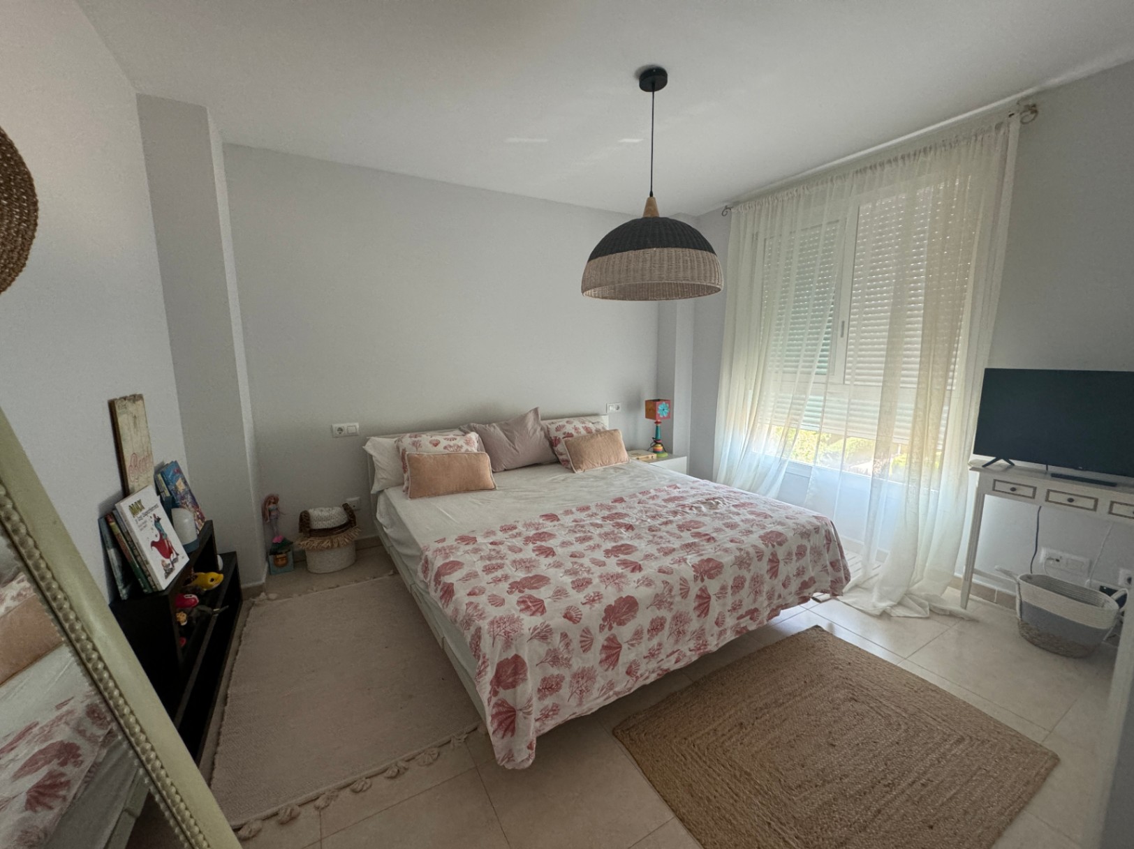 Helles Schlafzimmer mit Doppelbett, Textilien im Meeresdesign, einer Hängeleuchte aus Naturgeflecht und großem Fenster mit transparenten Vorhängen.