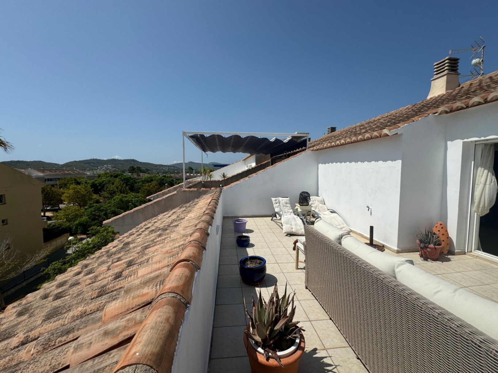 Langgestreckte Dachterrasse mit grauem Geflechtsofa, weißen Sonnenliegen und Terrakotta-Ziegeln, die einen weiten Blick auf die umliegenden grünen Hügel bietet.