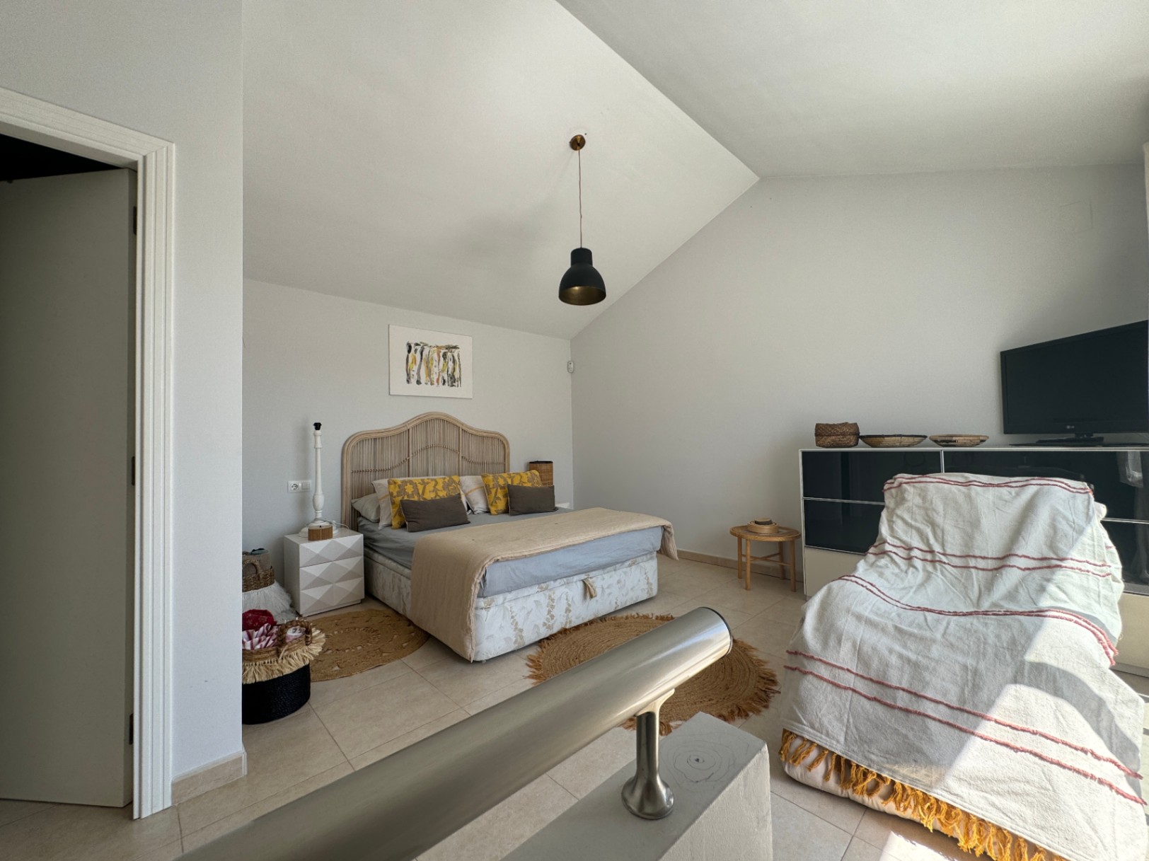 Schlafzimmer mit Dachschräge, Kopfteil aus Korbgeflecht und schwarzer Pendelleuchte. Fliesenboden, Jute-Teppiche und ein Metallgeländer im Vordergrund prägen den Raum.