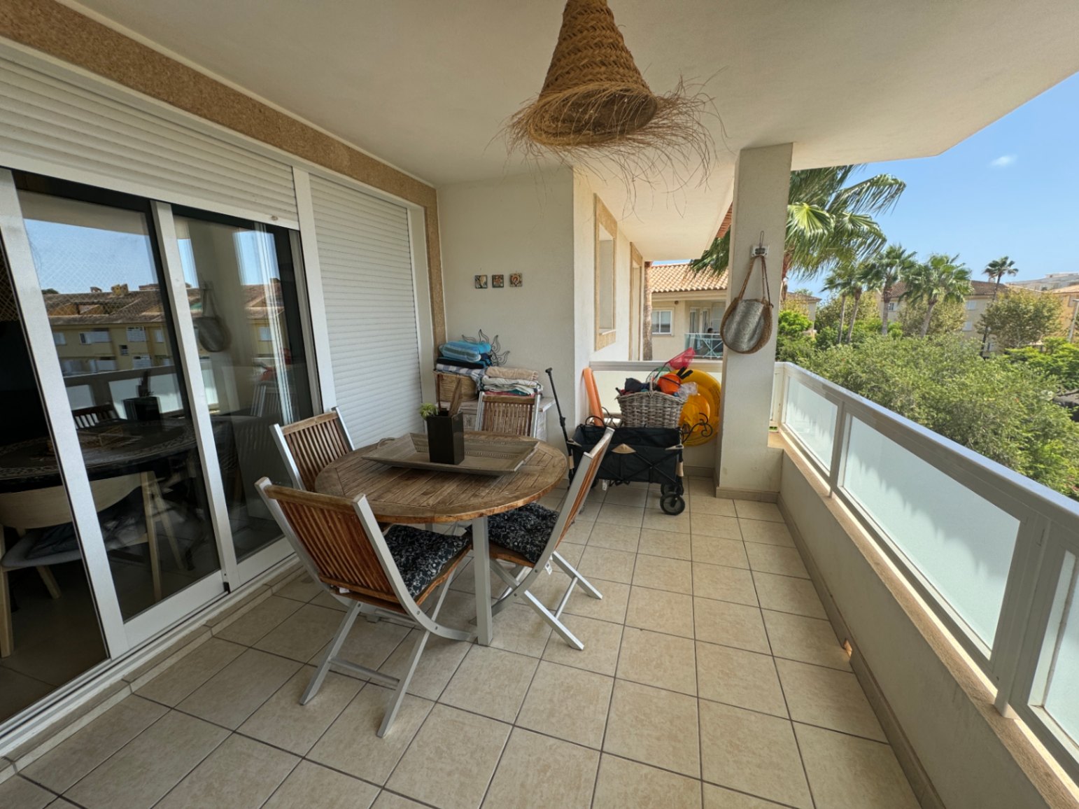 Überdachte Terrasse mit runder Holz-Sitzgruppe, Fliesenboden und Glasgeländer mit Blick auf Palmen und die umliegende Bebauung.