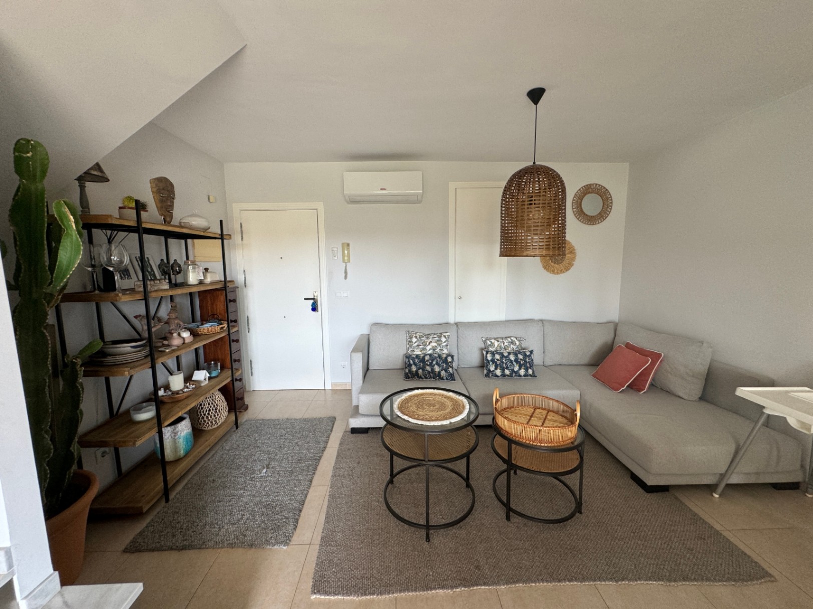 Wohnbereich mit grauem Ecksofa, Rattan-Pendelleuchte und industriellem Holzregal. Helle Fliesenböden und natürliche Dekorelemente prägen das moderne Ambiente.