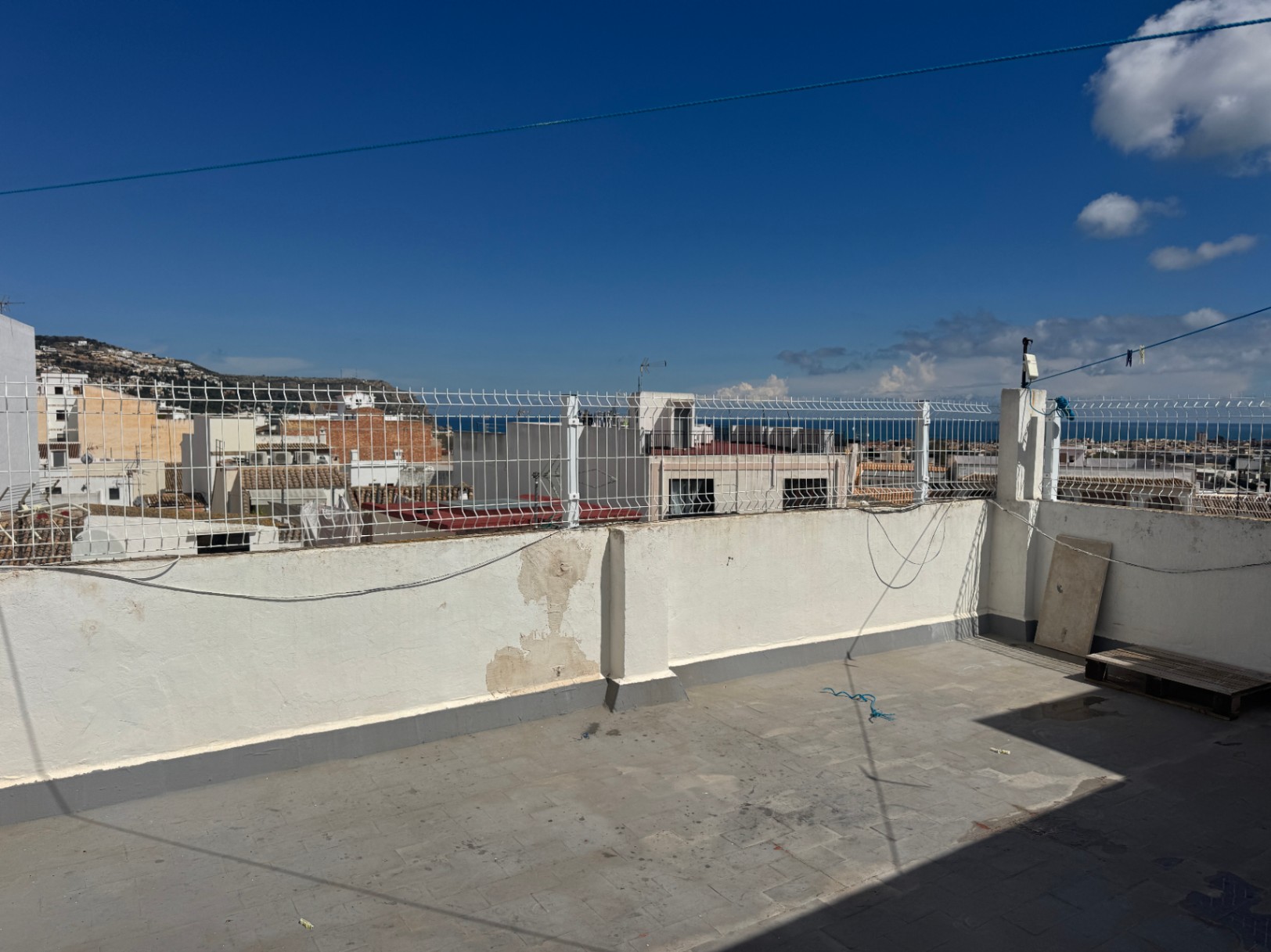 Dachterrasse mit Fliesenboden und weißen Umfassungsmauern mit Drahtzaun, mit Blick auf die Stadt, die umliegenden Hügel und das Meer.