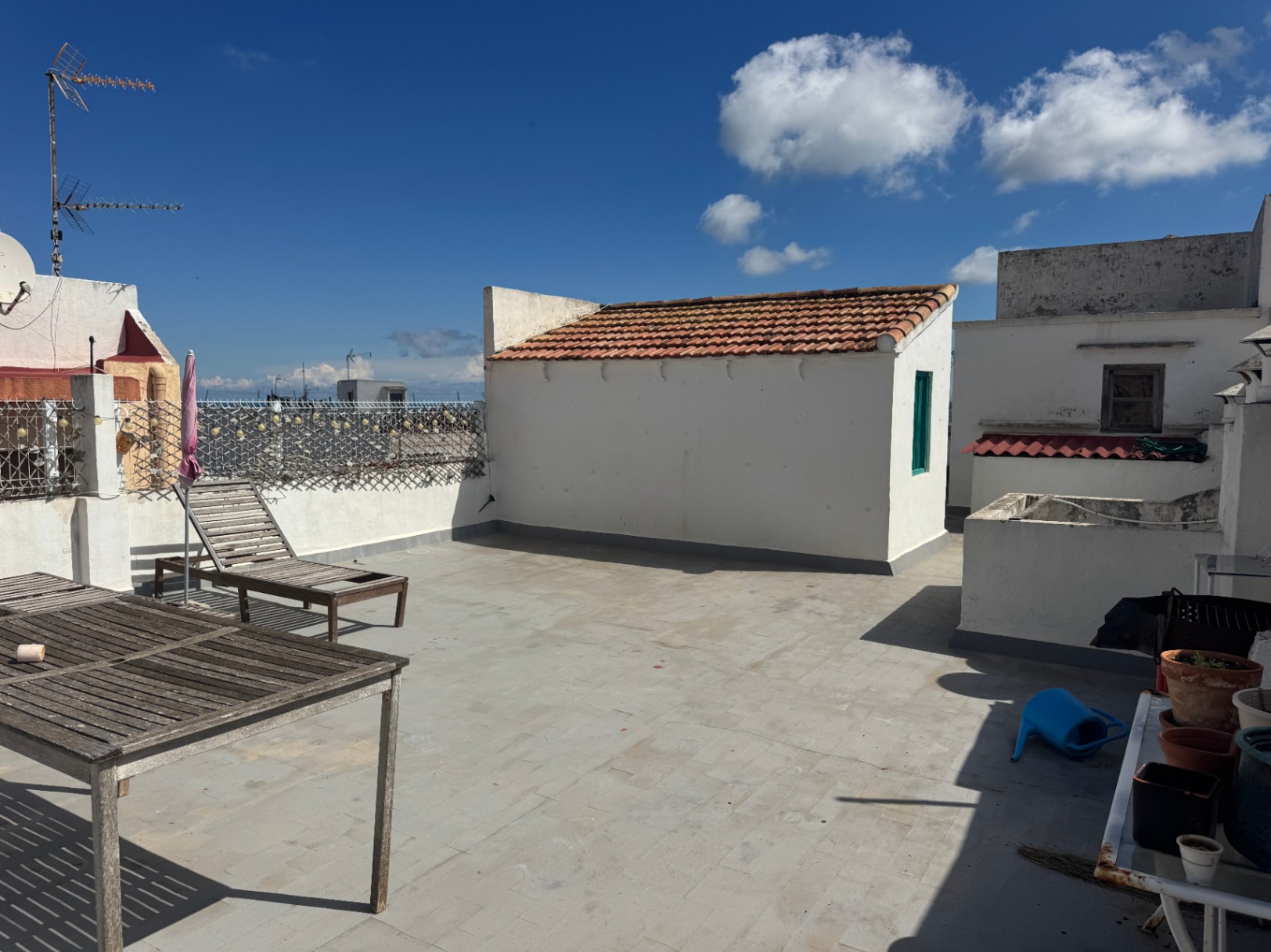 Dachterrasse mit Fliesenboden, verwitterten Holzmöbeln und einem weißen Nebengebäude mit traditionellem Ziegeldach unter blauem Himmel.