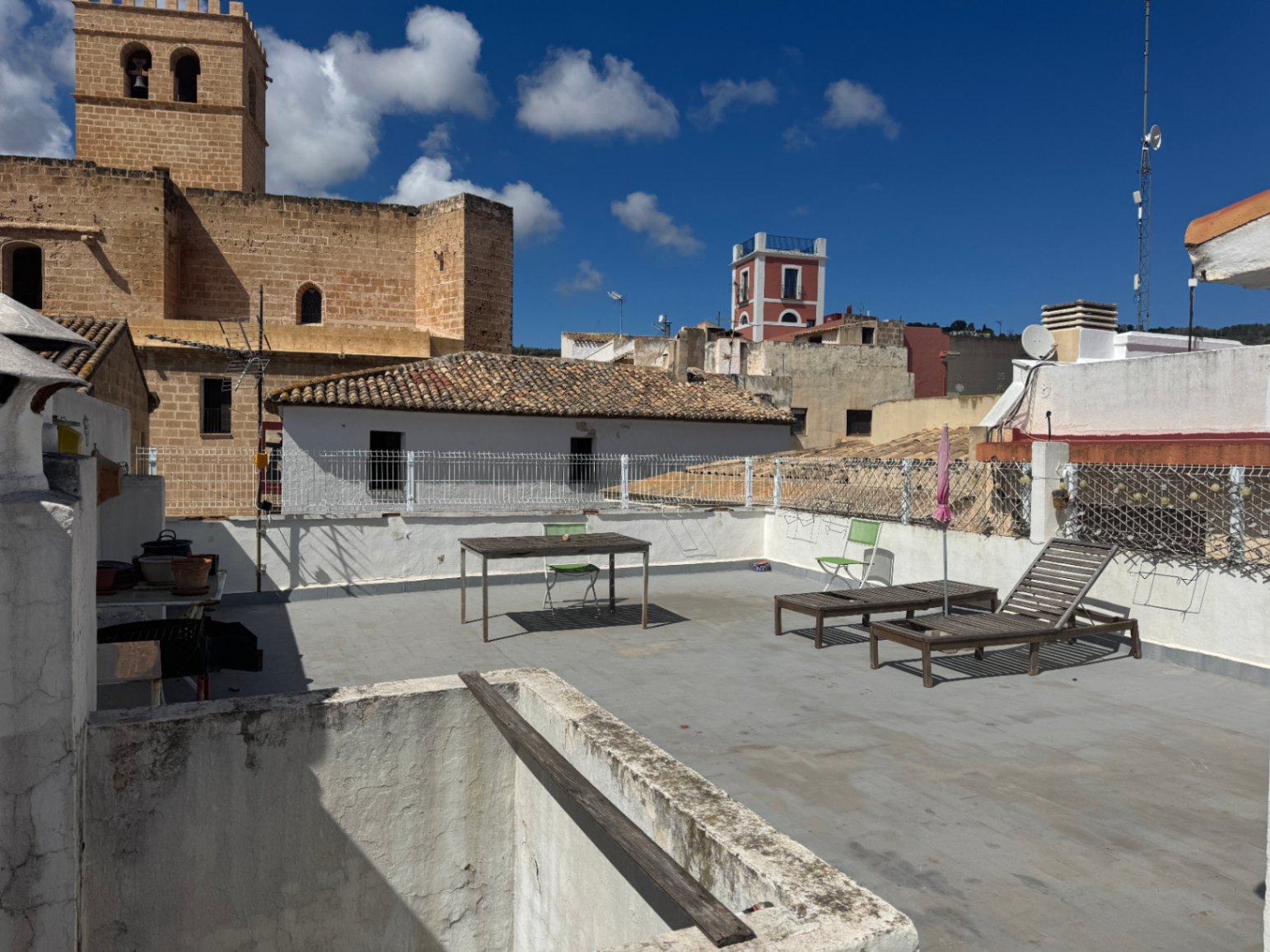 Dachterrasse mit hölzernen Sonnenliegen, Esstisch und Blick auf einen historischen Steinturm sowie die umliegenden Dächer des Dorfes.