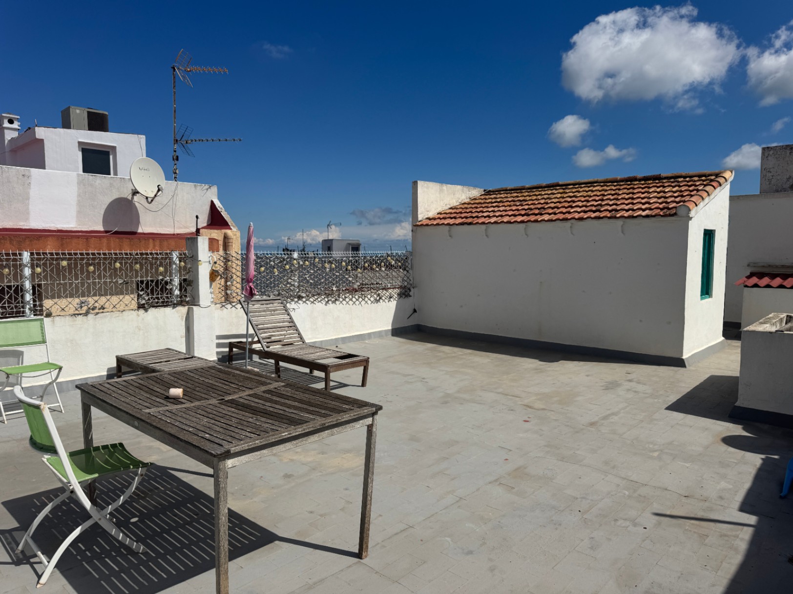 Großzügige Dachterrasse mit grauen Bodenfliesen, verwitterten Holzmöbeln und weißen Außenwänden unter strahlend blauem Himmel.