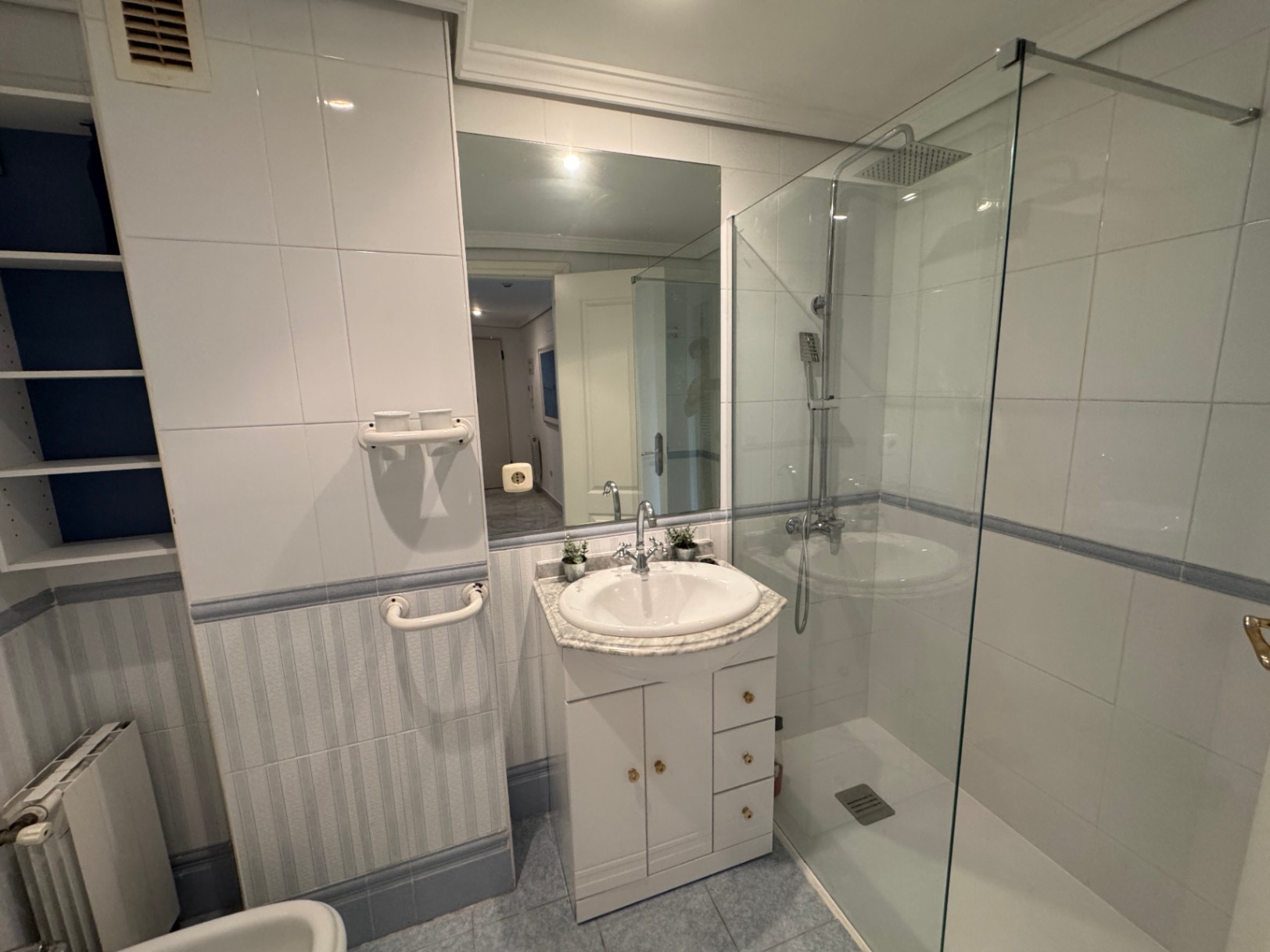 Badezimmer mit bodengleicher Glasdusche, weißem Waschtisch mit Marmorplatte und integrierten Regalen. Die Wände sind mit weißen und gestreiften Fliesen gestaltet.